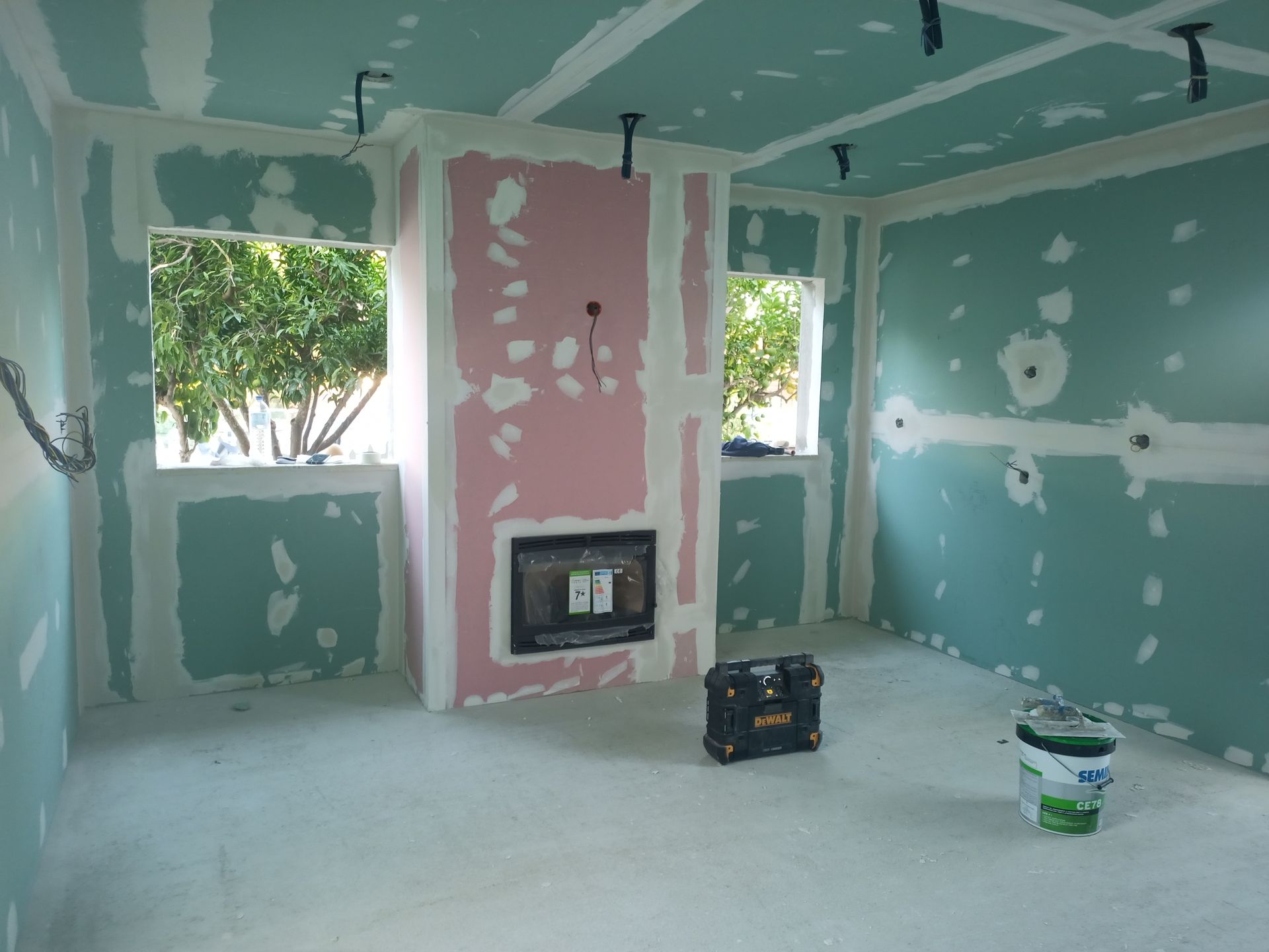 Interior de um cômodo em construção; drywall, lareira, janelas, fiação elétrica, ferramentas, balde de tinta.