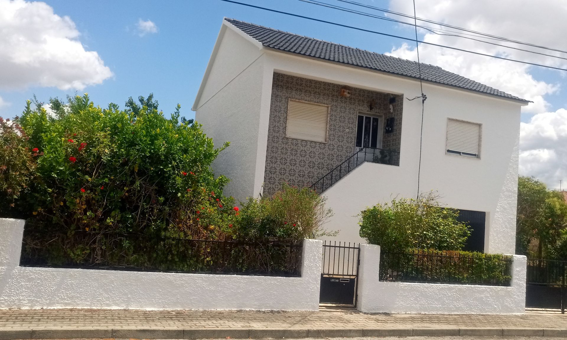 Casa branca de dois andares com telhado cinza escuro e fachada de pedra texturizada, cercada por vegetação e um muro branco.