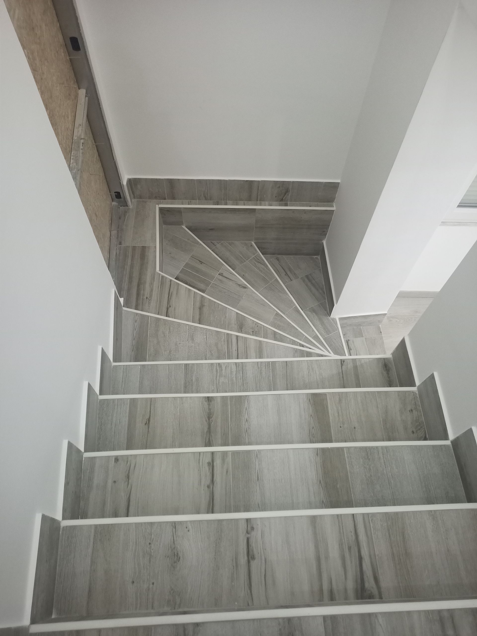 Escada com degraus revestidos com azulejos imitando madeira cinza e acabamento branco, que desce a partir de um patamar.