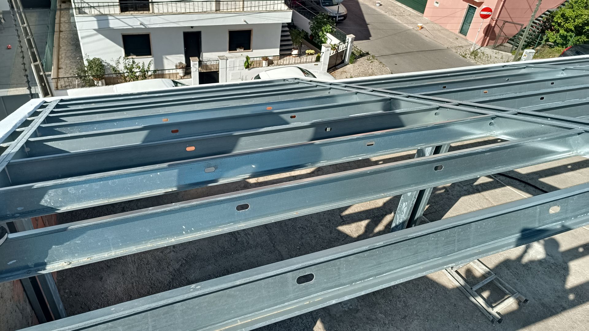 Estrutura metálica de um telhado em construção, vista externamente, com um prédio e uma rua visíveis ao fundo.