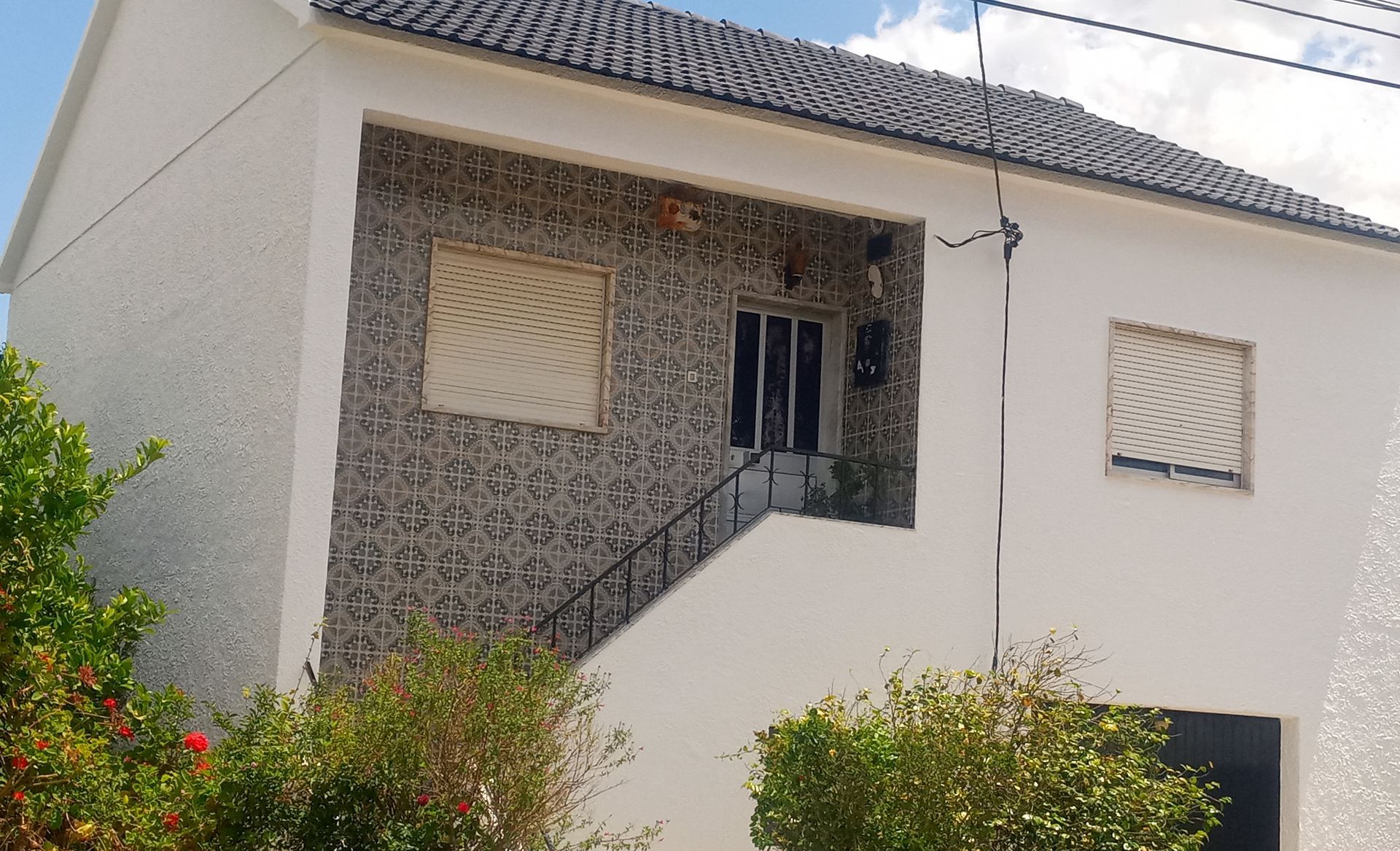 Casa branca de dois andares com fachada de azulejos cinza na entrada e janelas fechadas com venezianas. Telhado preto, céu azul.