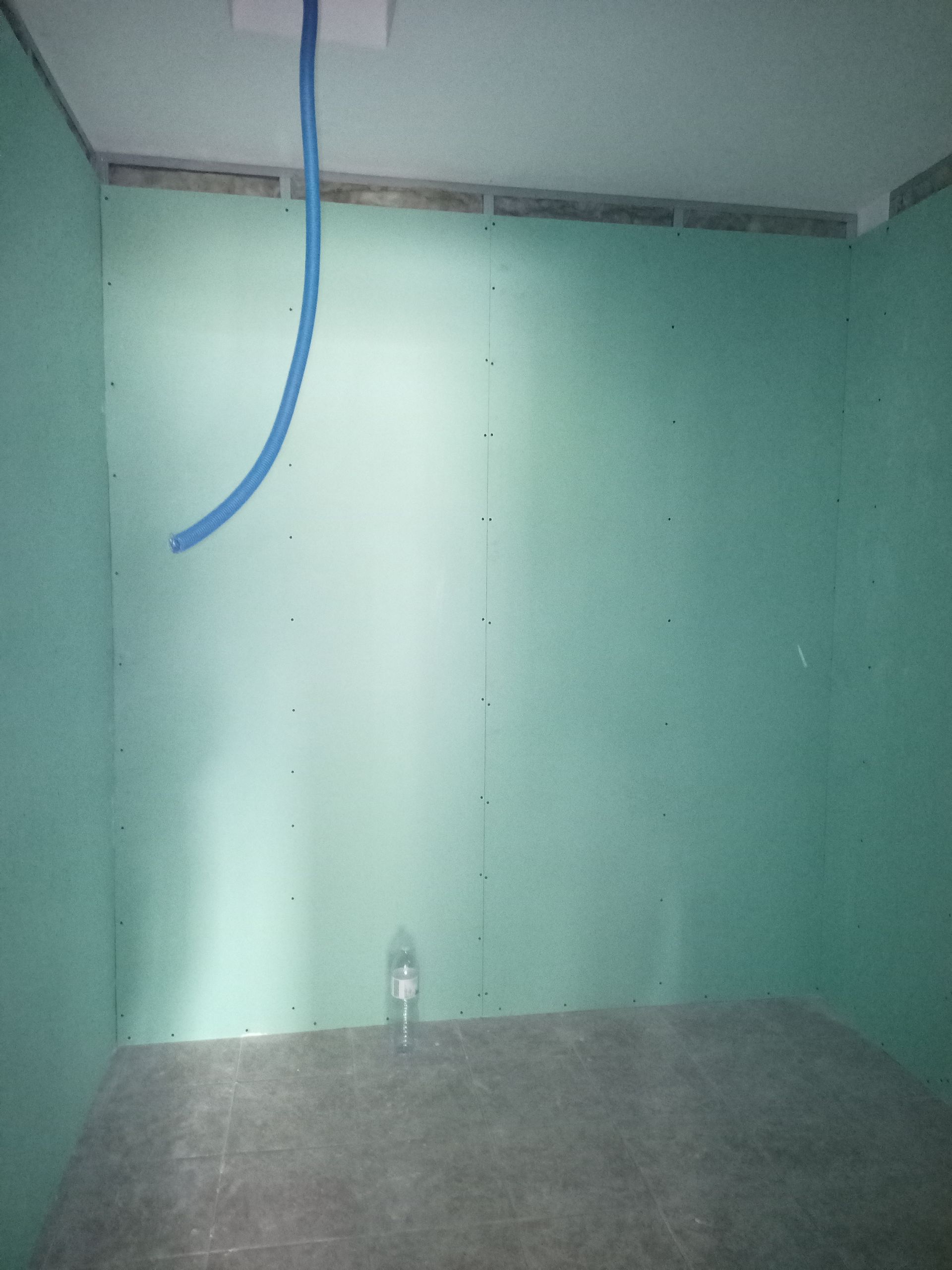Quarto em construção com paredes verdes, com um cano azul pendurado e uma garrafa no chão.