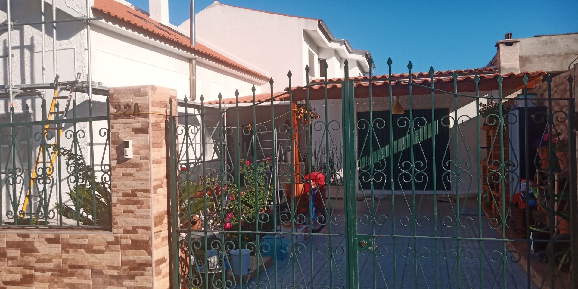 Casa com portão de ferro e telhado de telha vermelha, com plantas e colunas de tijolos, sob um céu azul.
