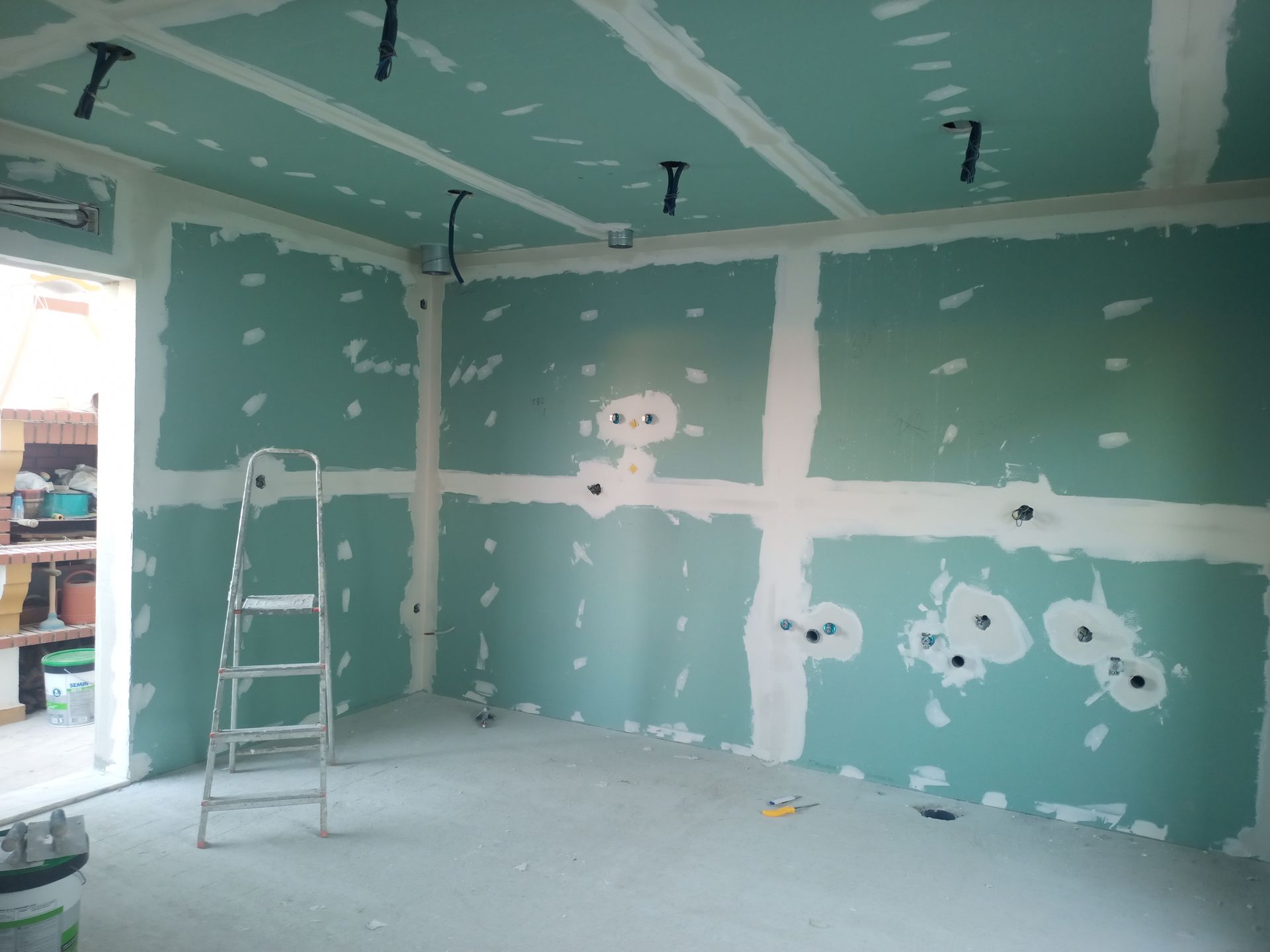 Interior de um cômodo em construção, com drywall verde, fita adesiva branca e uma escada. A fiação elétrica sai do teto.