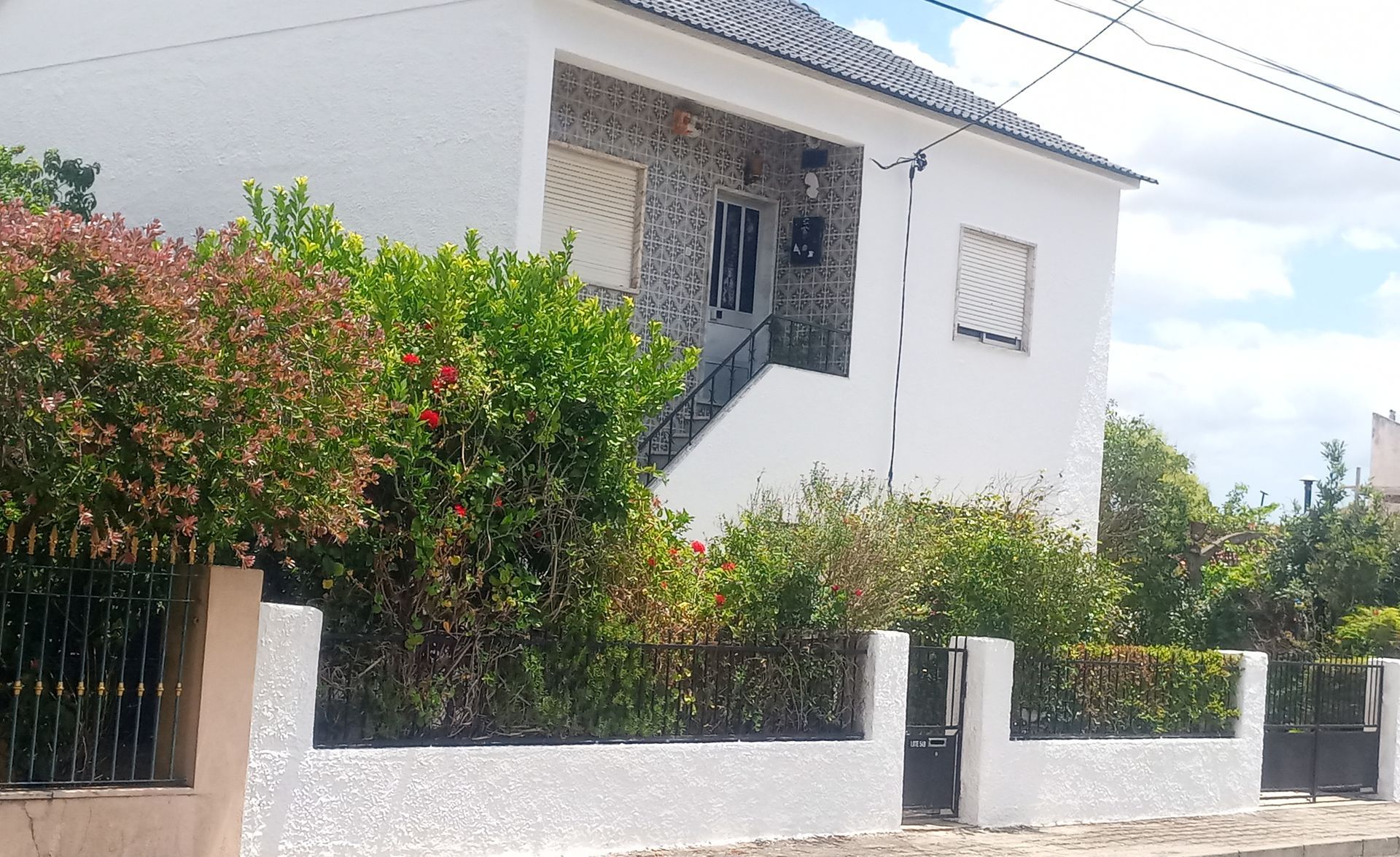 Casa branca de dois andares com fachada de pedra, cercada por arbustos e uma cerca de metal preta em um dia ensolarado.