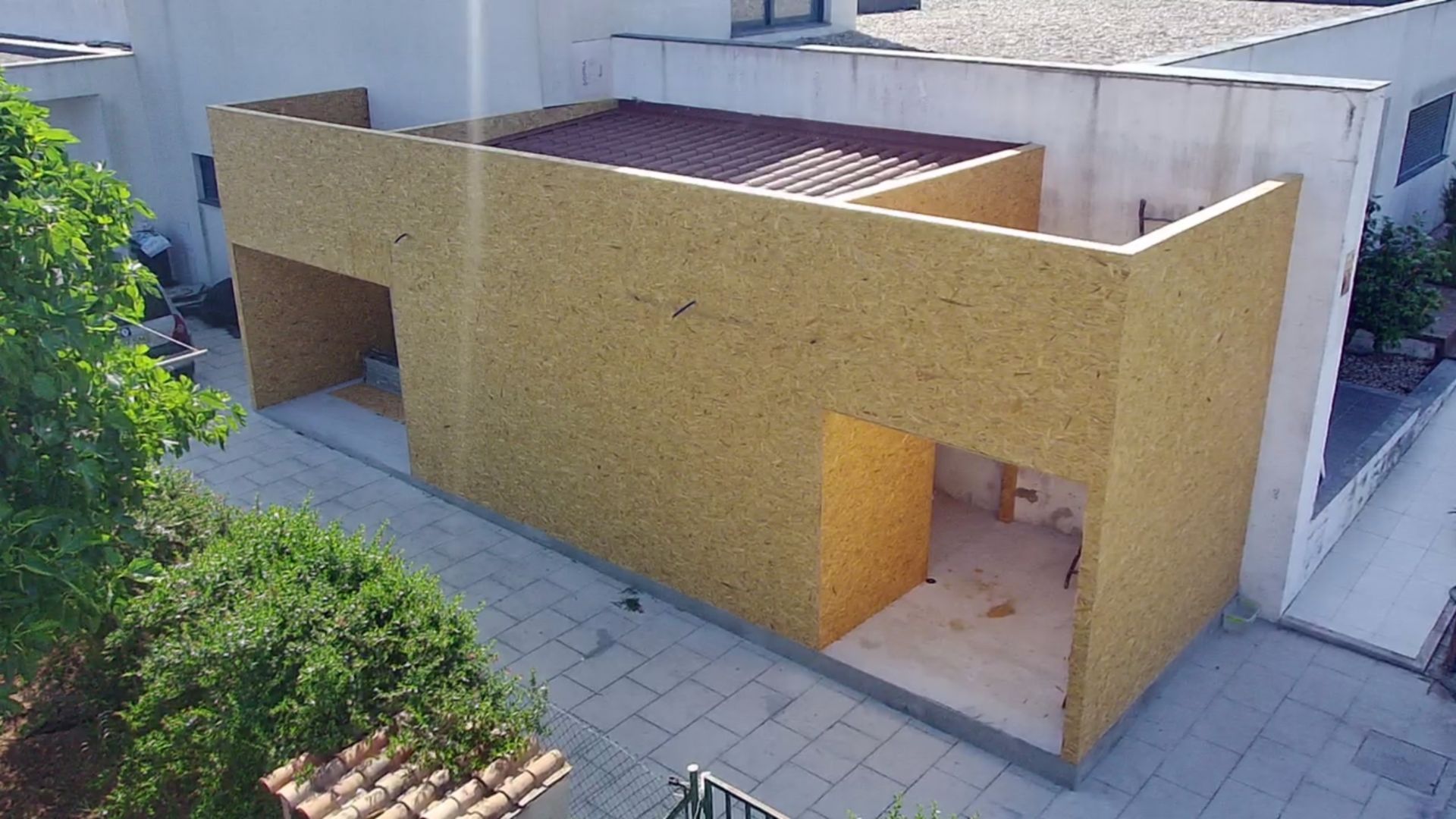 Vista exterior de um edifício em construção, com paredes revestidas de painéis de madeira clara. Telhado vermelho, portas abertas e um pátio pavimentado.