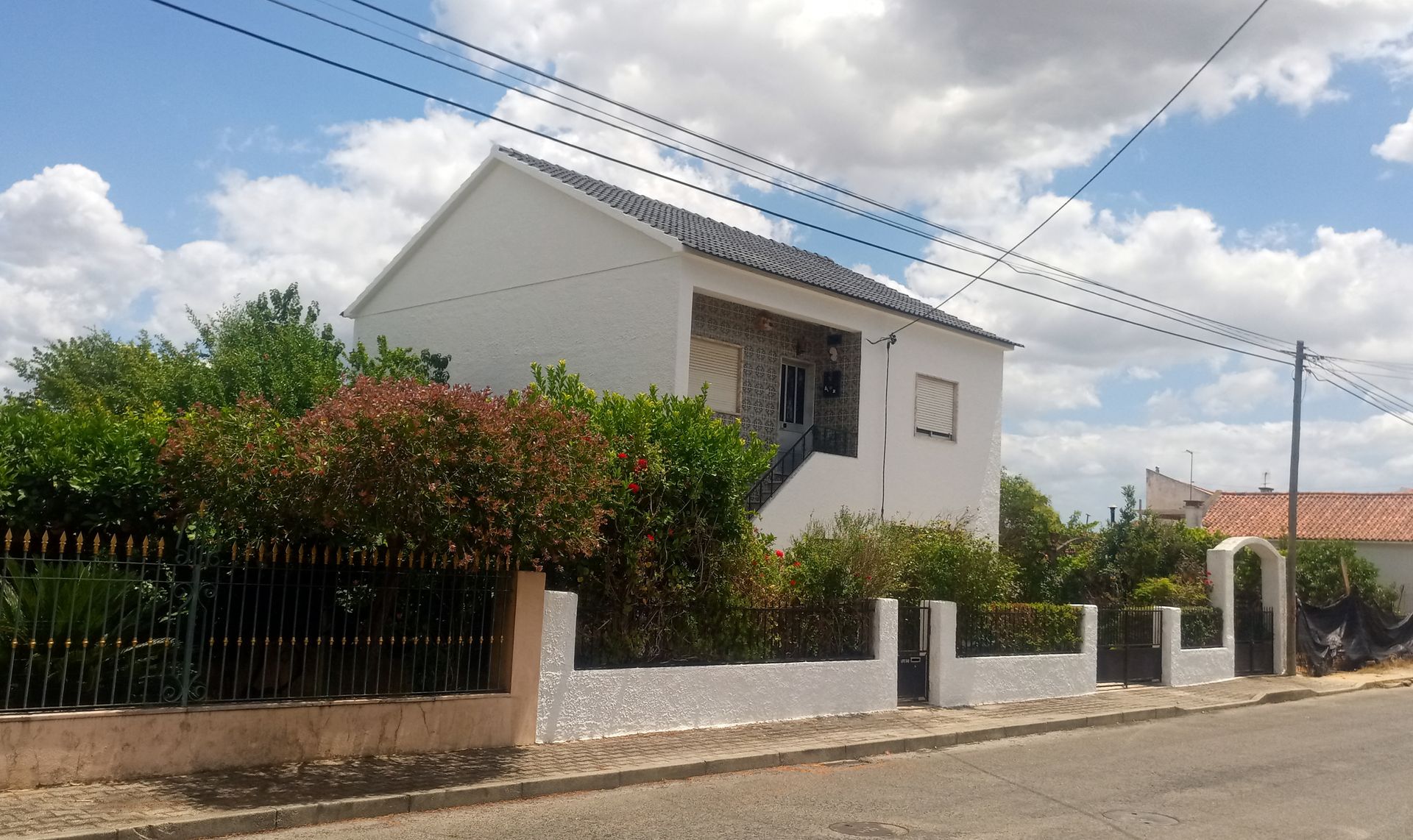 Casa branca de dois andares com uma pequena varanda e um jardim, atrás de uma cerca baixa, sob um céu nublado.