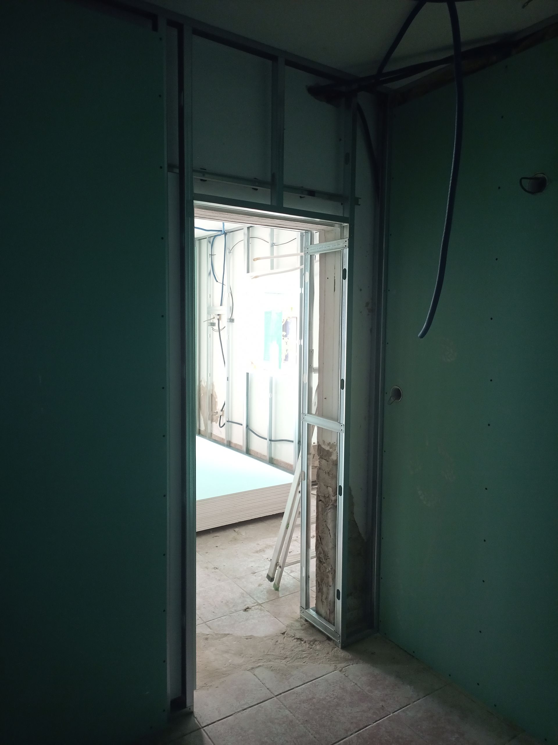 Vista interna da entrada de um cômodo em construção; paredes de gesso e estrutura metálica visíveis, paredes verdes, luz proveniente do cômodo adjacente.