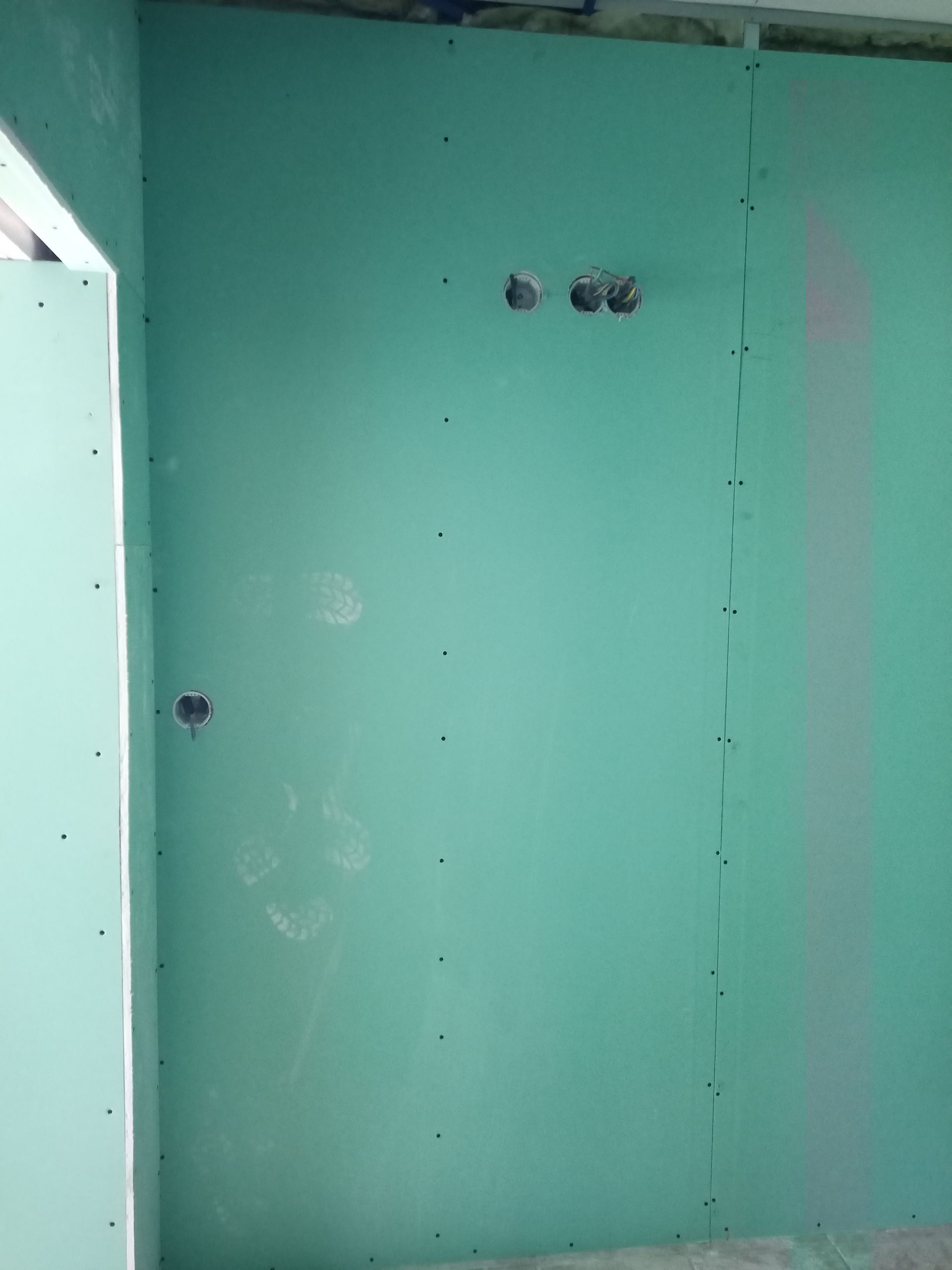 Interior com paredes de gesso cartonado verde e tomadas elétricas, sem acabamento.