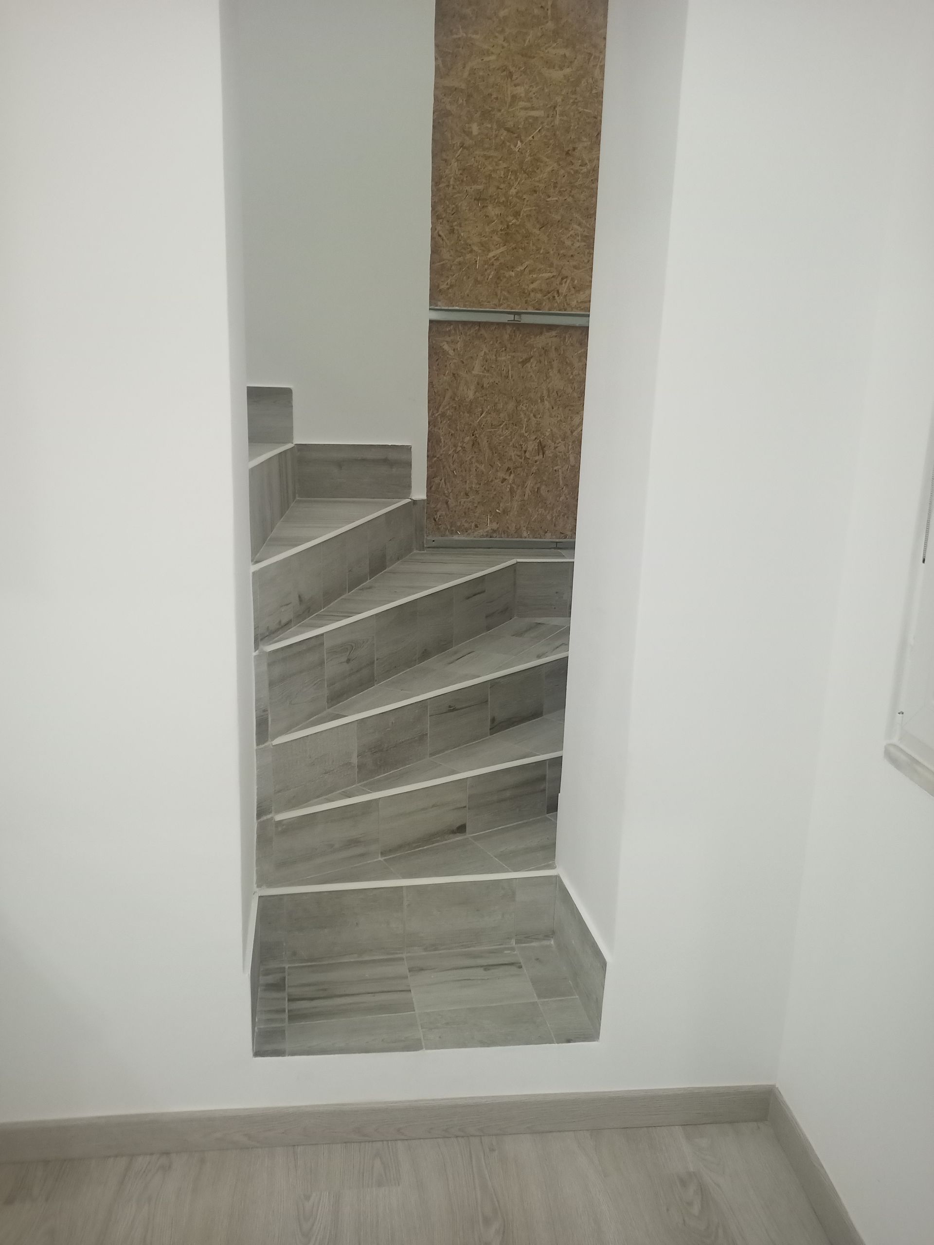 Escada em um espaço estreito com paredes brancas, degraus cinza e painéis de madeira na lateral.