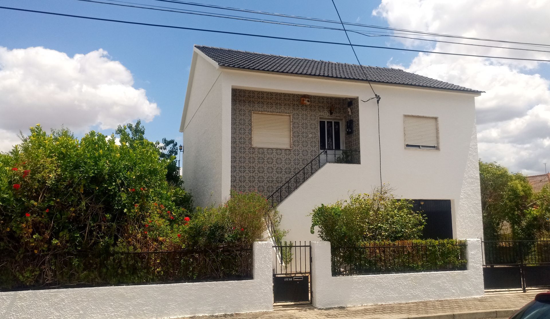 Casa branca de dois andares com telhado de telhas escuras, venezianas cinza e portão de metal preto. Arbustos verdes na frente.