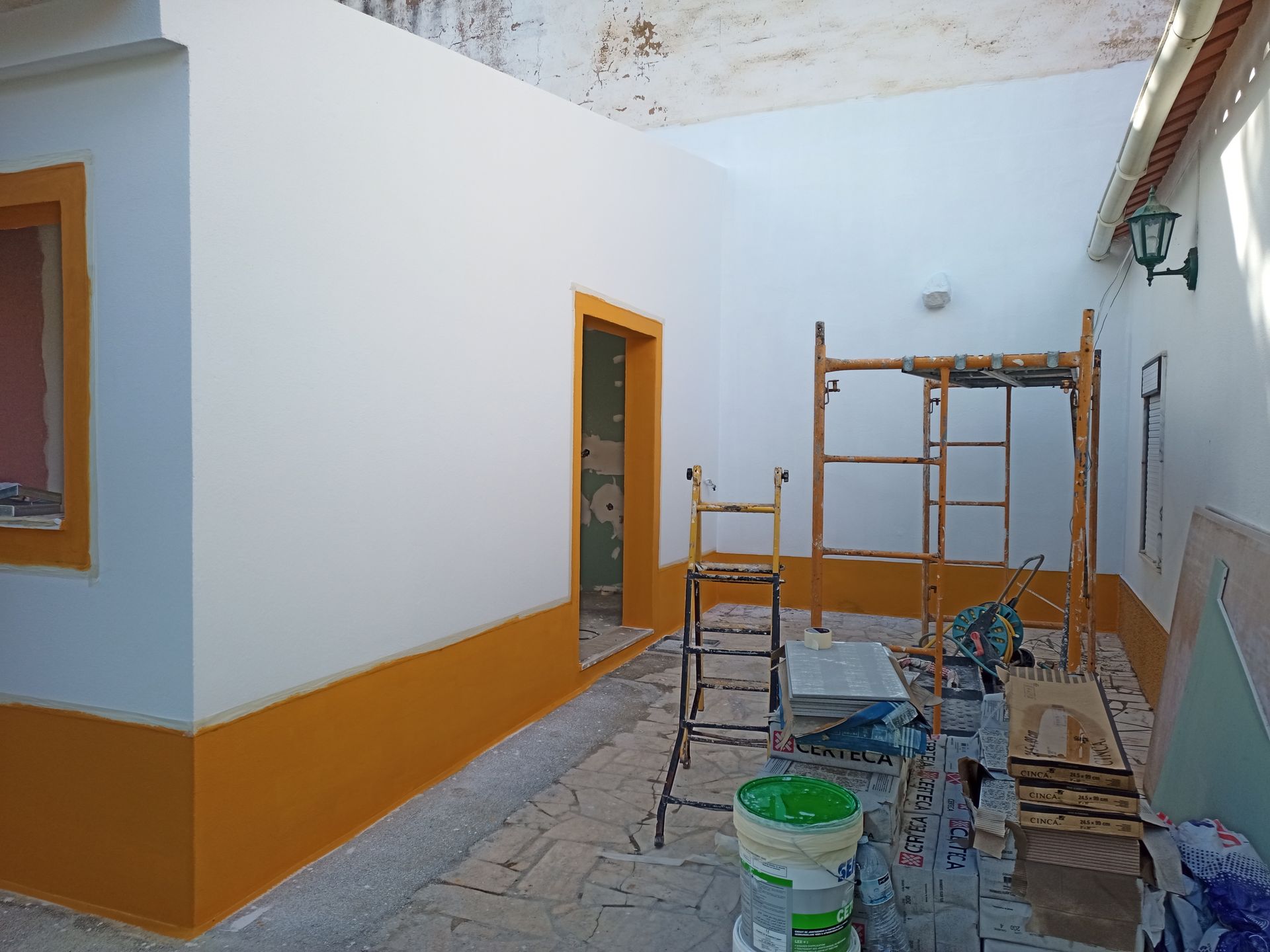 Quarto em reforma; paredes brancas com detalhes em laranja, andaimes, ferramentas e porta aberta.