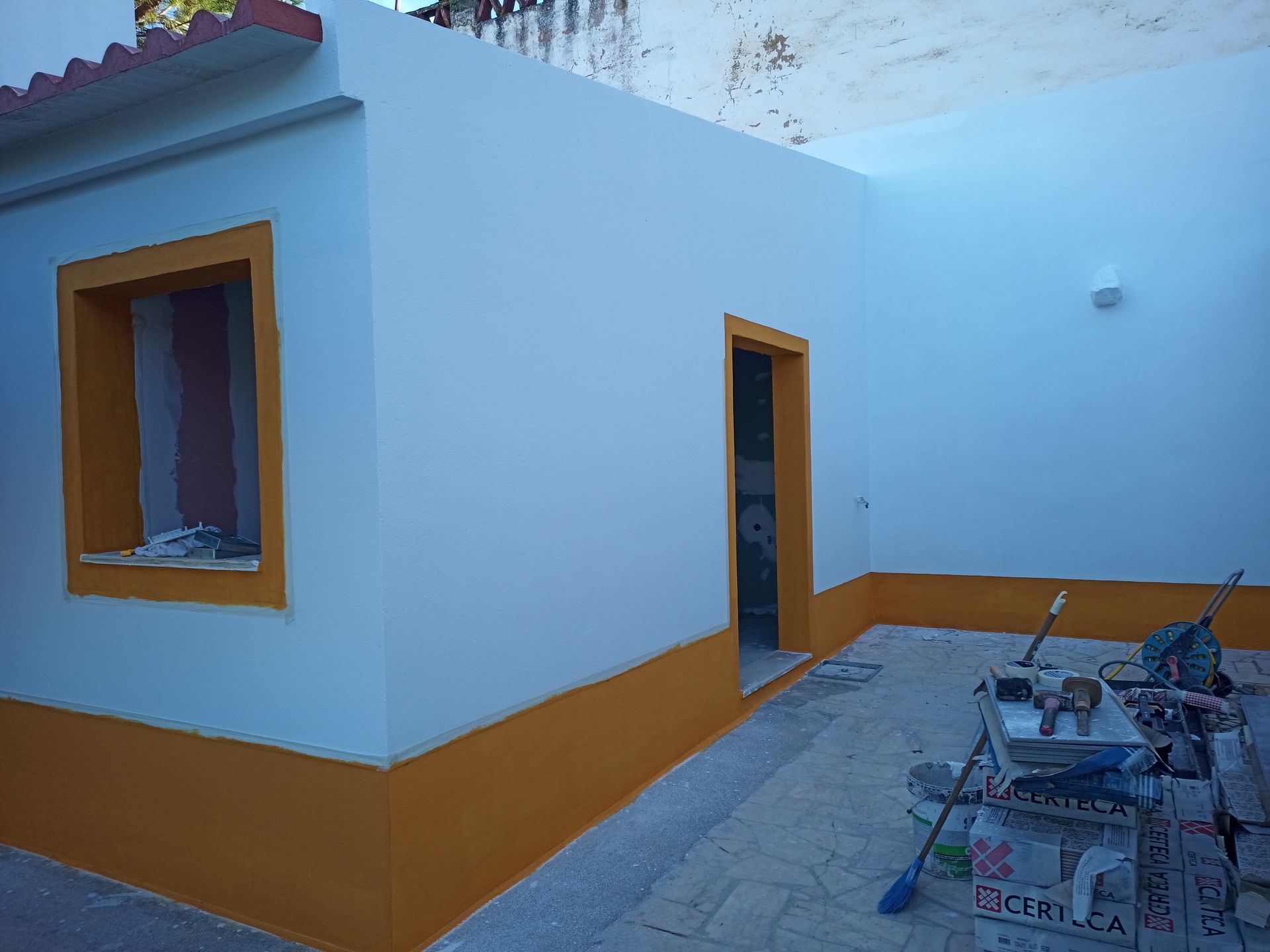 Exterior de um prédio branco com detalhes em laranja ao redor de uma janela e porta. Ferramentas estão no pátio.