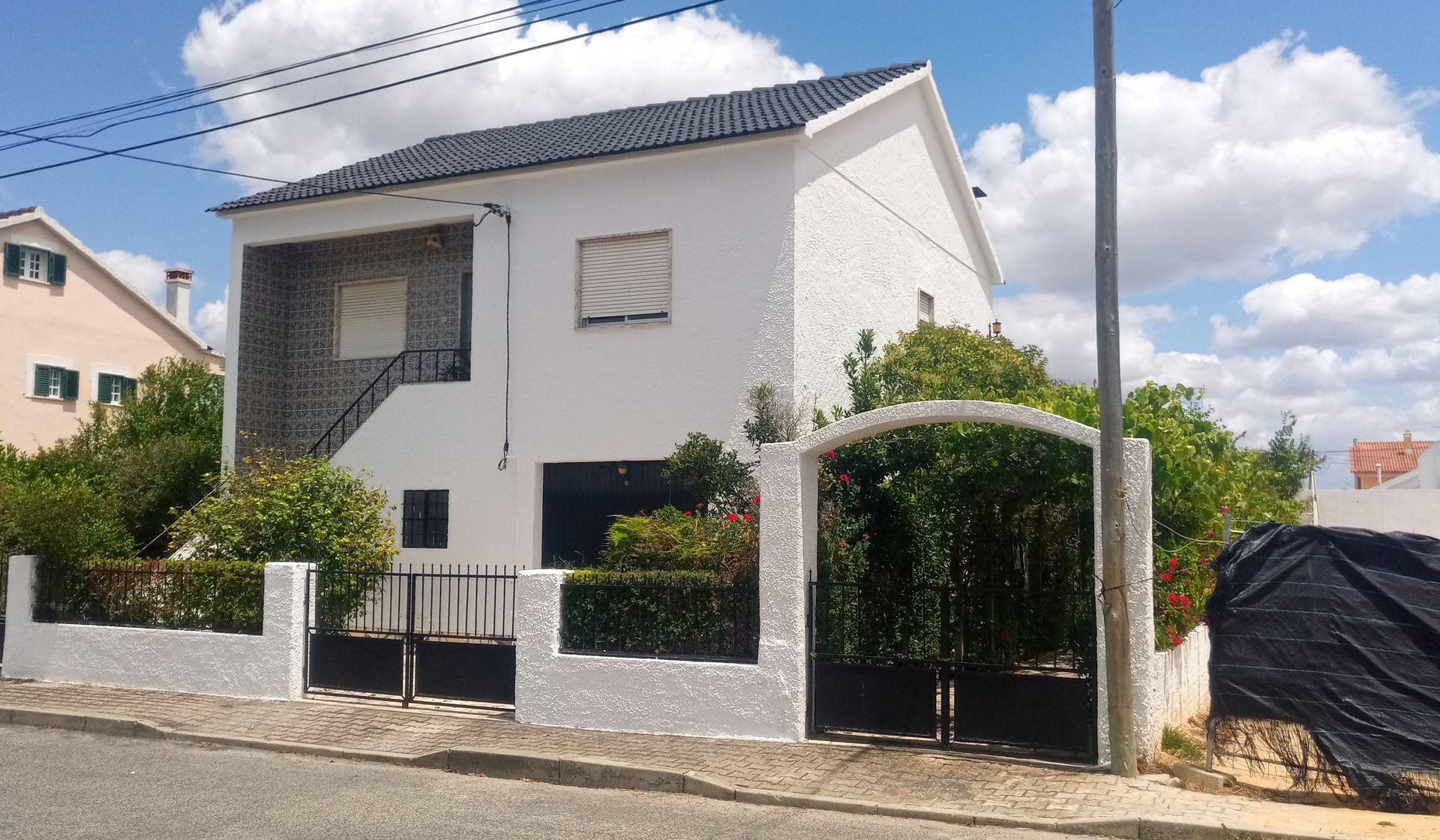 Casa branca de dois andares com telhado preto, pequeno jardim frontal, portão preto e vista para a rua com poste de luz.
