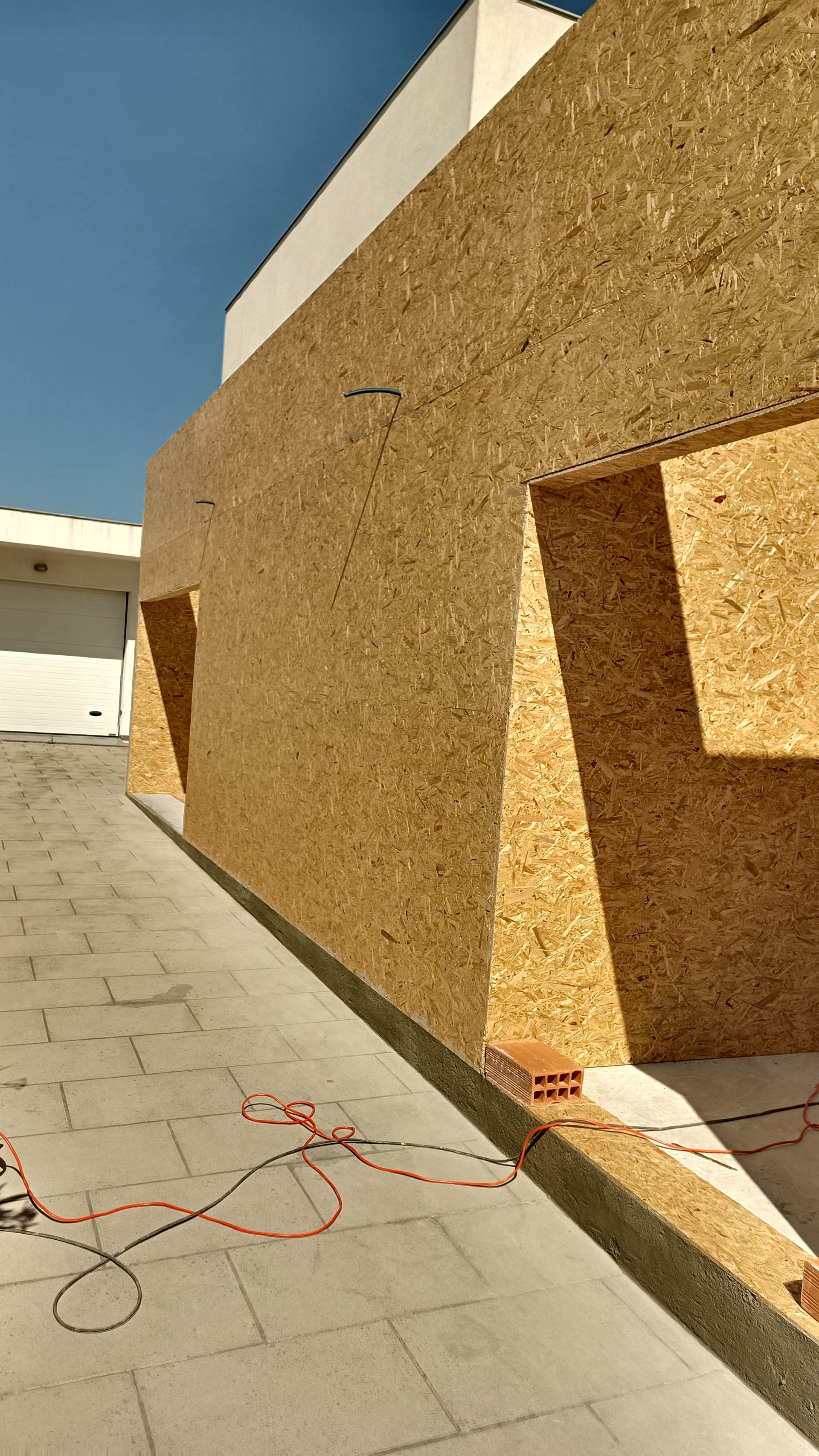 Exterior do edifício com painéis de madeira castanho-clara, duas janelas, pavimento cinzento, muita luz solar e fios elétricos.
