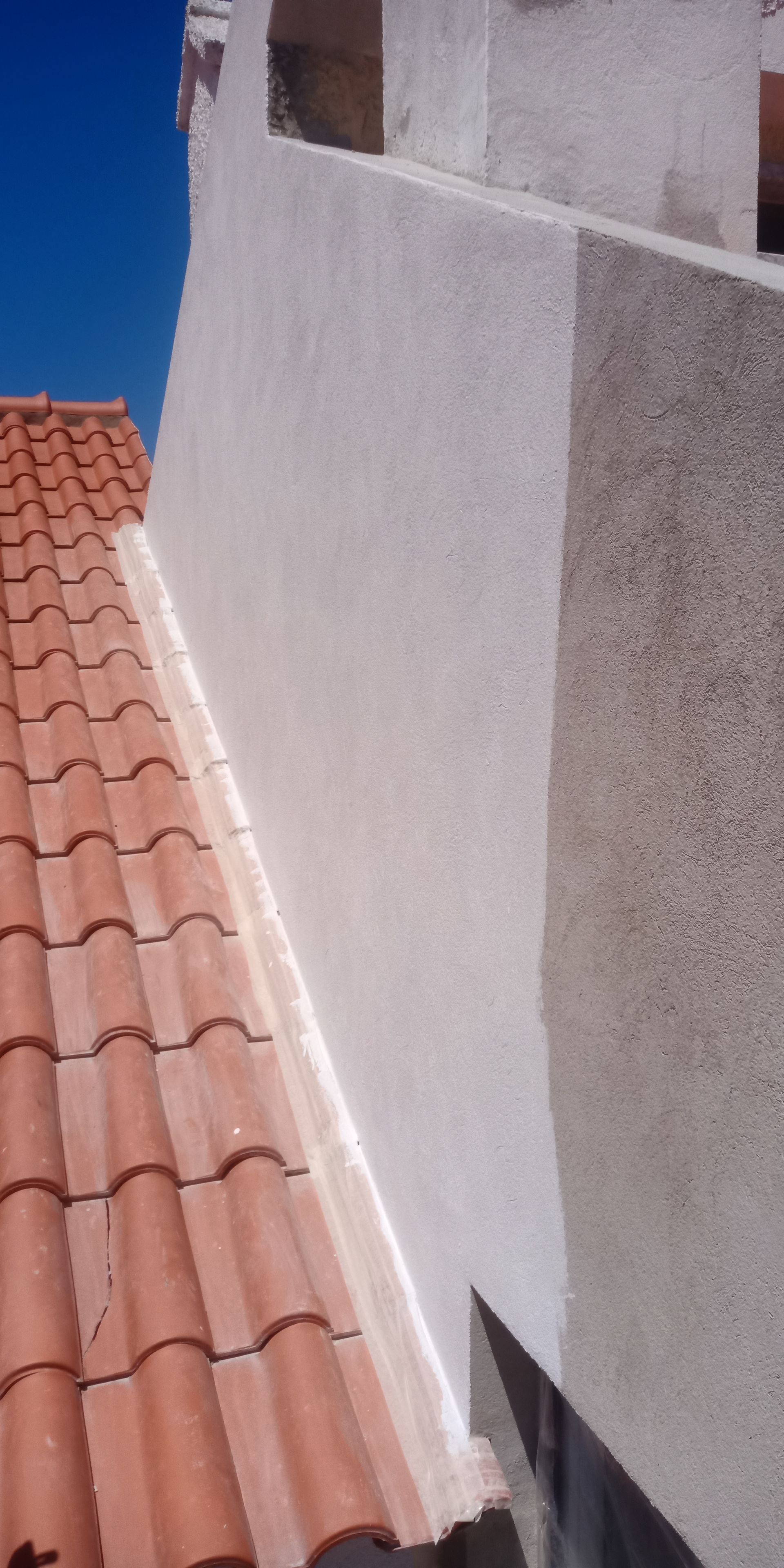 Telhado de telha vermelha encontra uma parede branca com textura de estuque, céu azul visível.