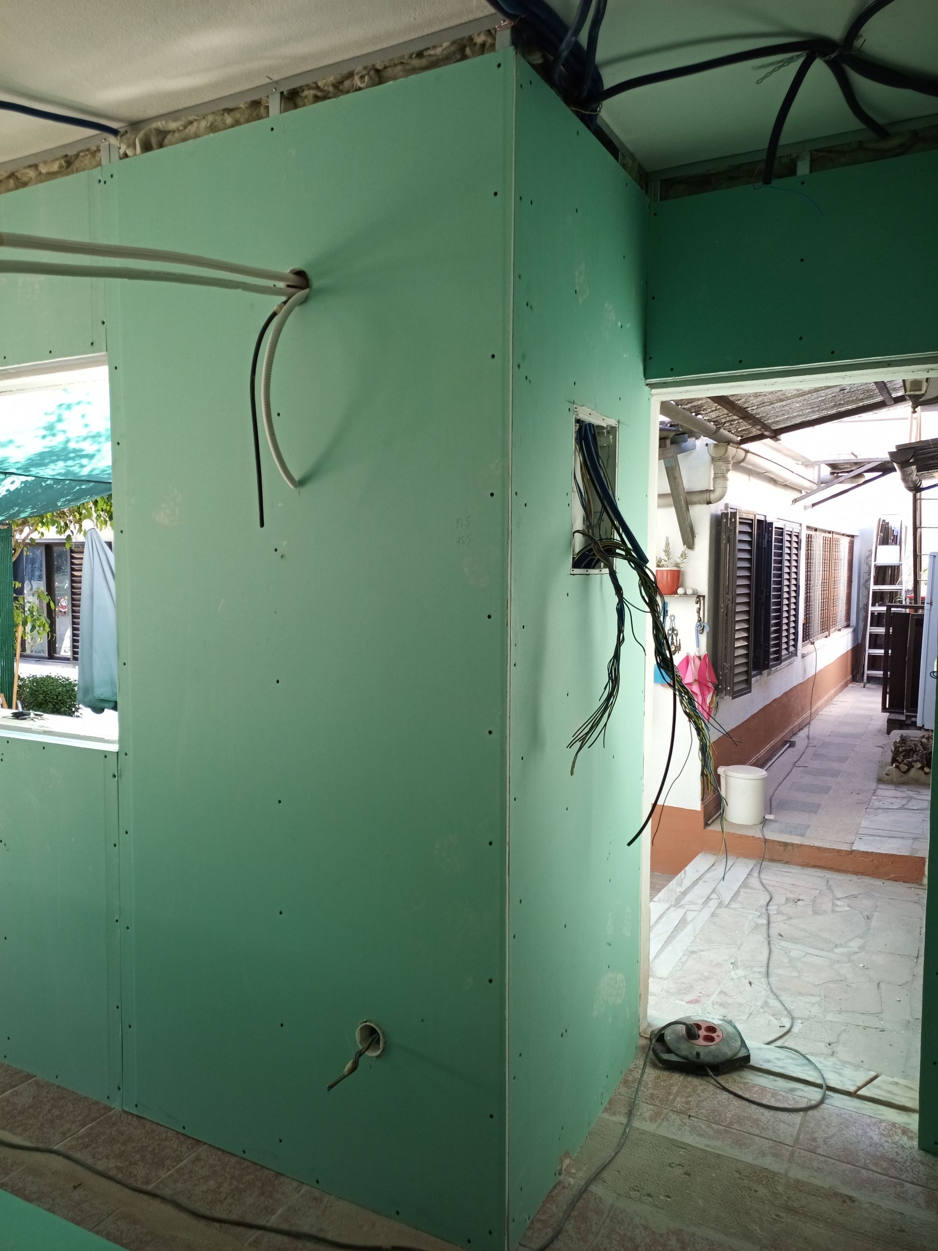 Vista interna: paredes de gesso cartonado verde em construção, fiação exposta, porta que dá acesso à área externa.