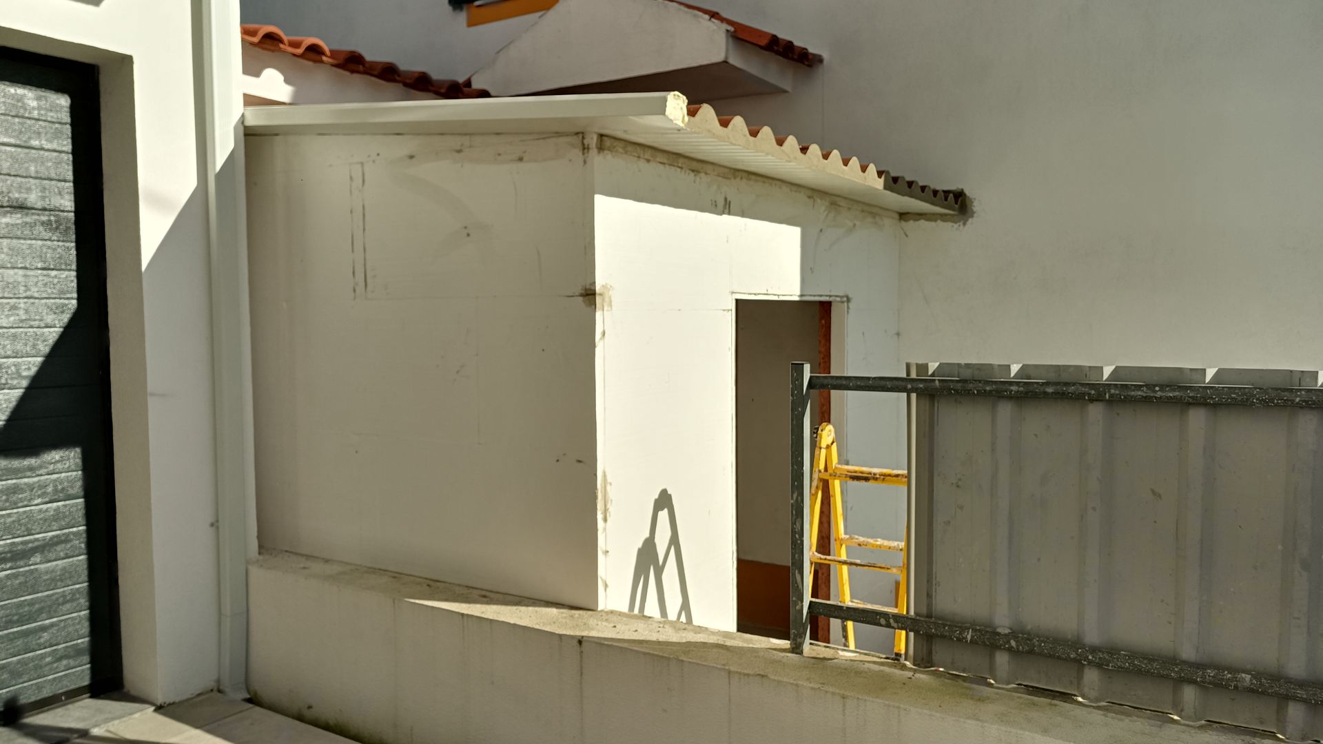Prédio branco com porta aberta, escada amarela e cerca de metal em uma rua ensolarada.