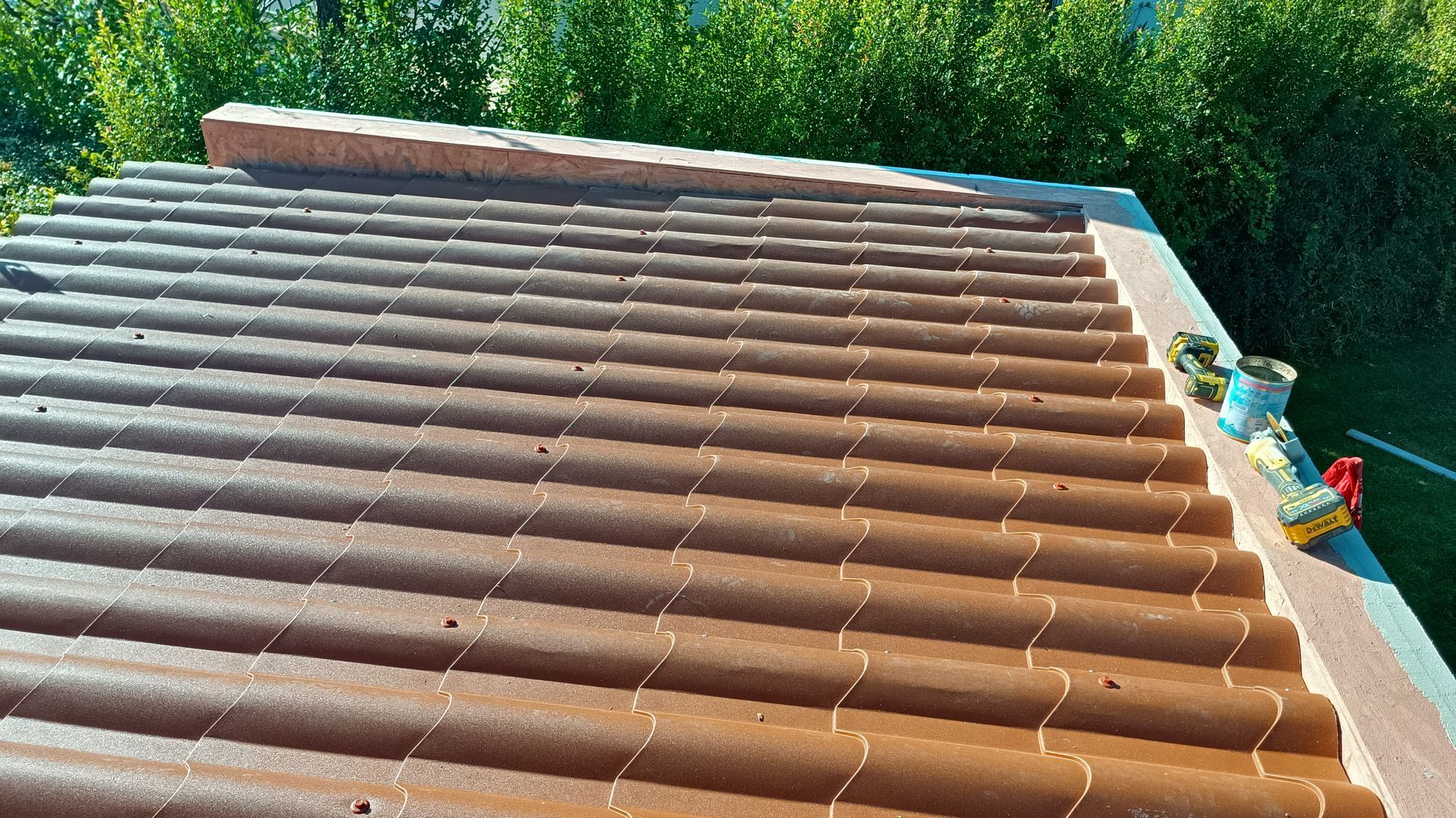 Telhado de telha marrom com ferramentas, visto de cima, folhagem verde ao fundo.