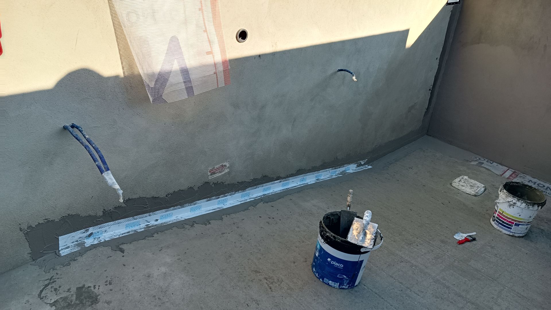 Canteiro de obras: paredes e piso de concreto cinza, encanamento e baldes com ferramentas.