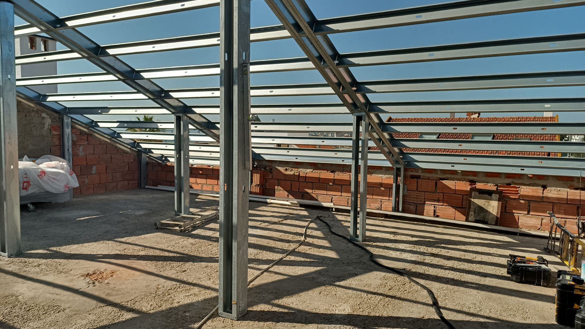 Vista interior de um edifício em construção, com vigas de aço, paredes de tijolo e luz solar visível.
