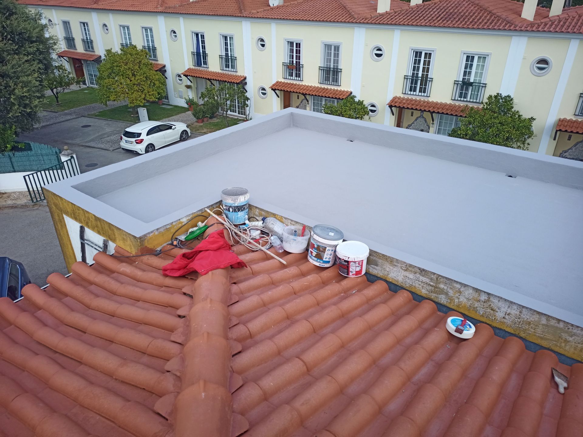 Um telhado plano pintado de cinza com latas de tinta e ferramentas em cima, ao lado de um telhado de telhas vermelhas e um prédio com varandas.