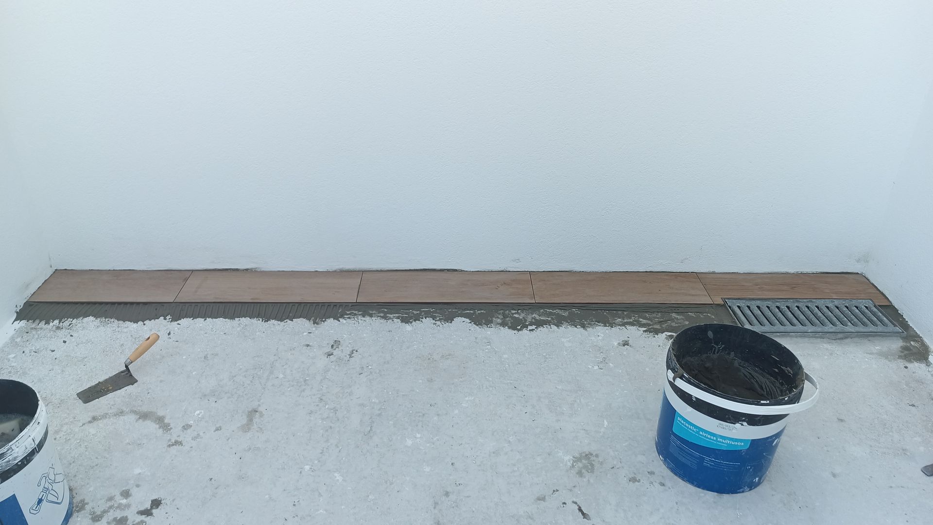 Construção de uma superfície revestida com azulejos em um espaço com paredes brancas e ralo. Baldes de material e uma espátula estão presentes.