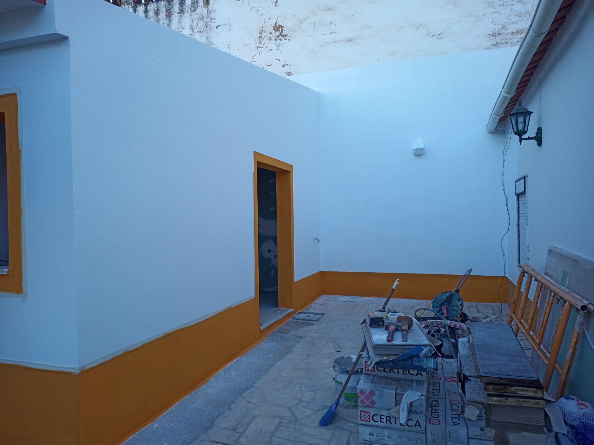 Exterior de um prédio com paredes brancas, detalhes em amarelo e porta aberta; ferramentas no chão.