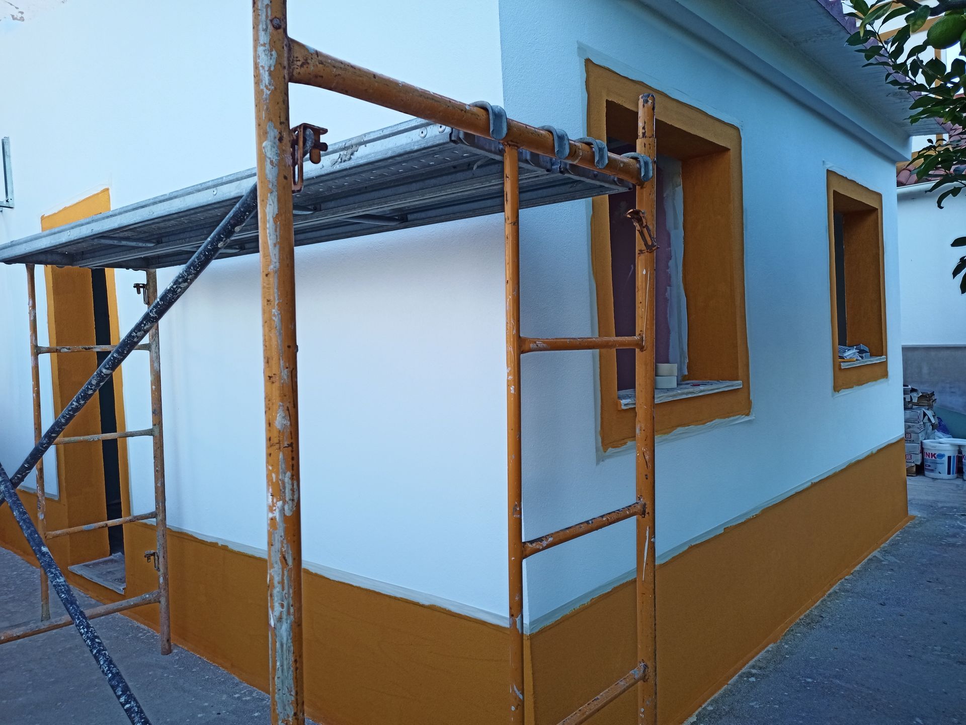 Exterior de uma casa com paredes brancas, detalhes em amarelo e andaimes.