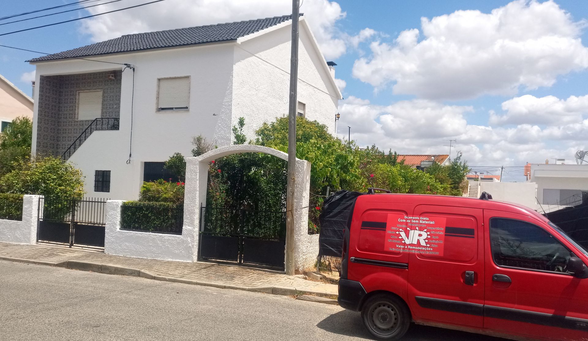 Casa branca de dois andares com telhado e portão escuros, van vermelha estacionada na rua.