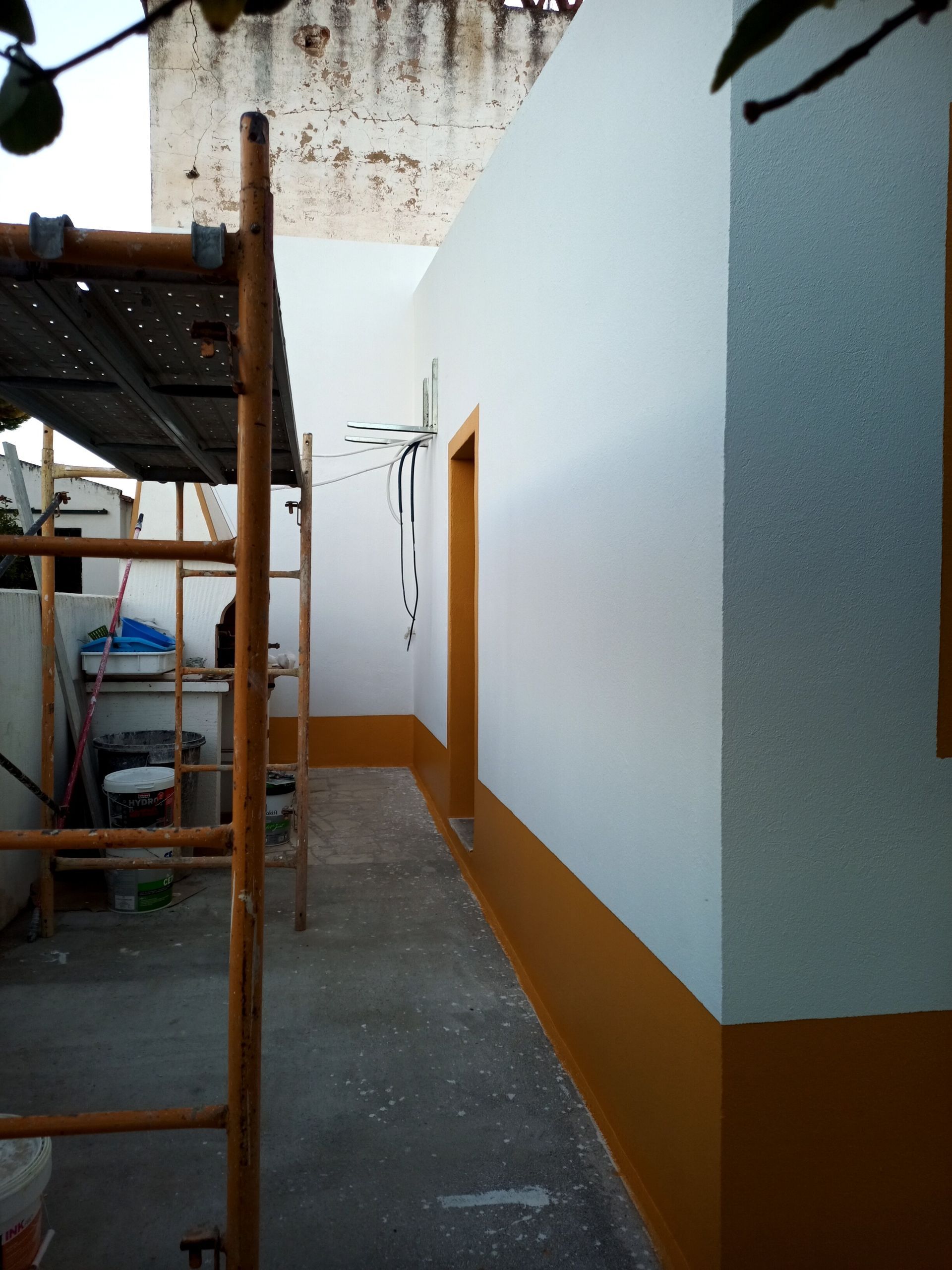 Exterior de um edifício com paredes brancas, base amarelo-mostarda e batente de porta. Andaimes à esquerda.
