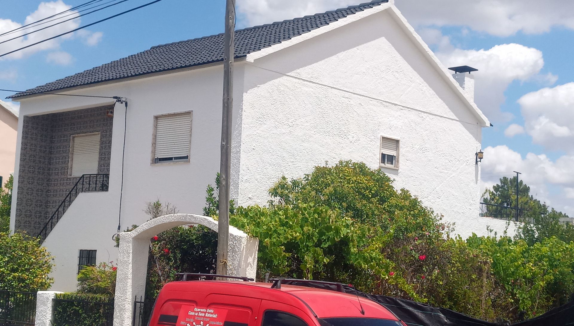 Casa branca com telhado preto, janelas pequenas, arbustos e um veículo vermelho em primeiro plano.