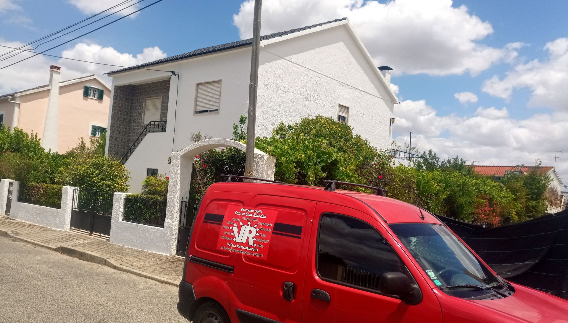 Uma carrinha vermelha estacionada na rua em frente a uma casa branca de dois andares com um portão. Céu azul e arbustos.