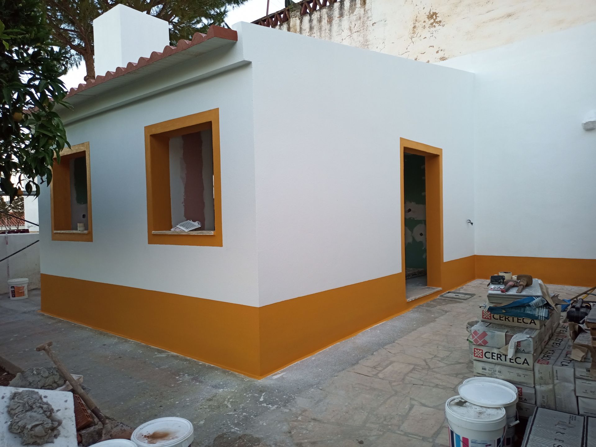 Exterior branco e amarelo de um pequeno prédio em construção; janelas e caixilhos de portas inacabados.