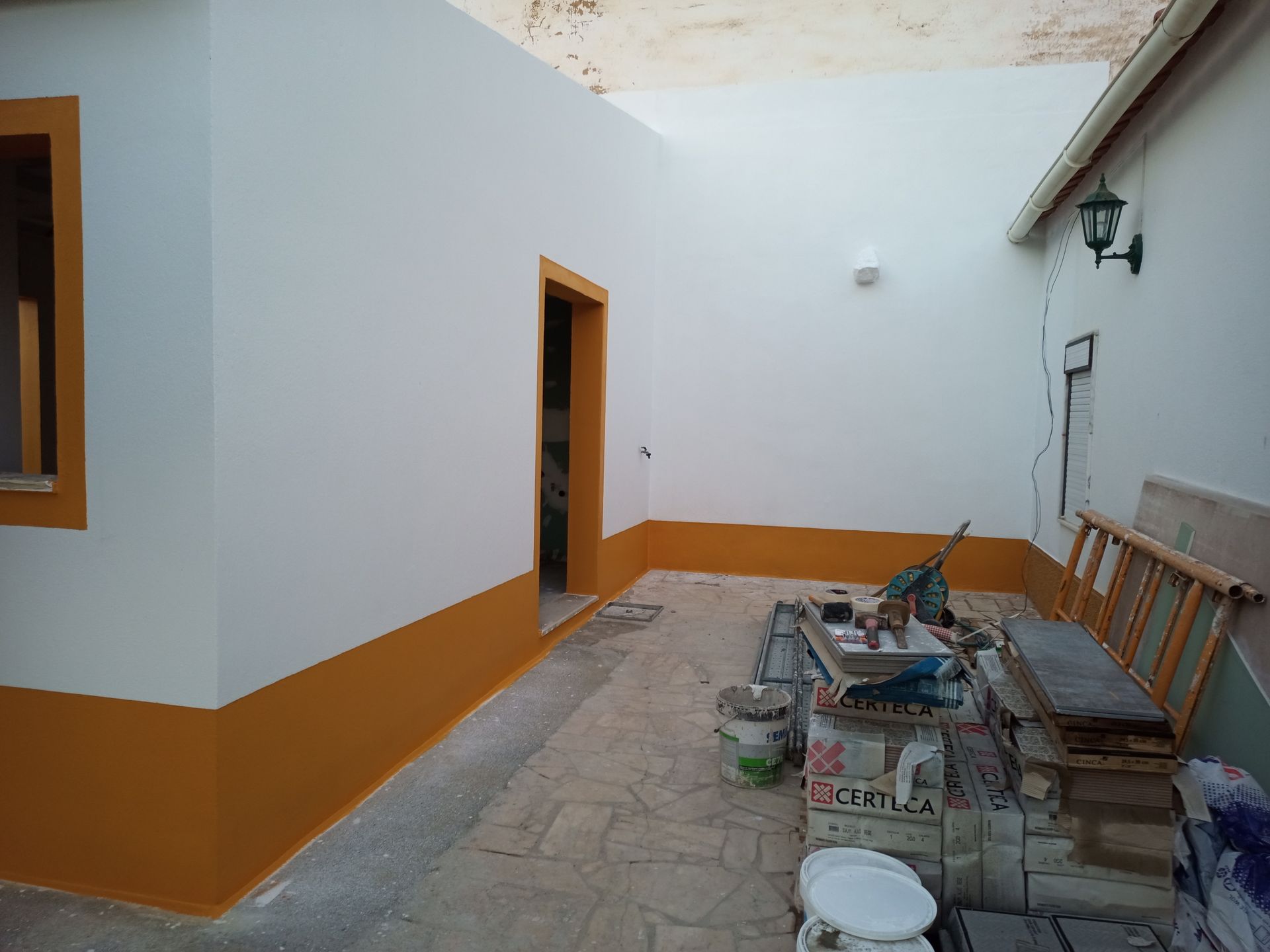 Exterior de um edifício com paredes pintadas de branco e laranja, e um pátio de pedra. Materiais de construção estão espalhados pelo local.