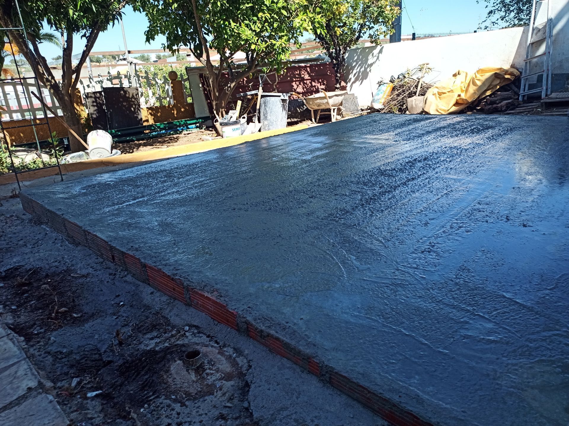 Laje de concreto recém-lançada, com fôrmas, em um quintal sob um céu azul, refletindo a luz do sol.