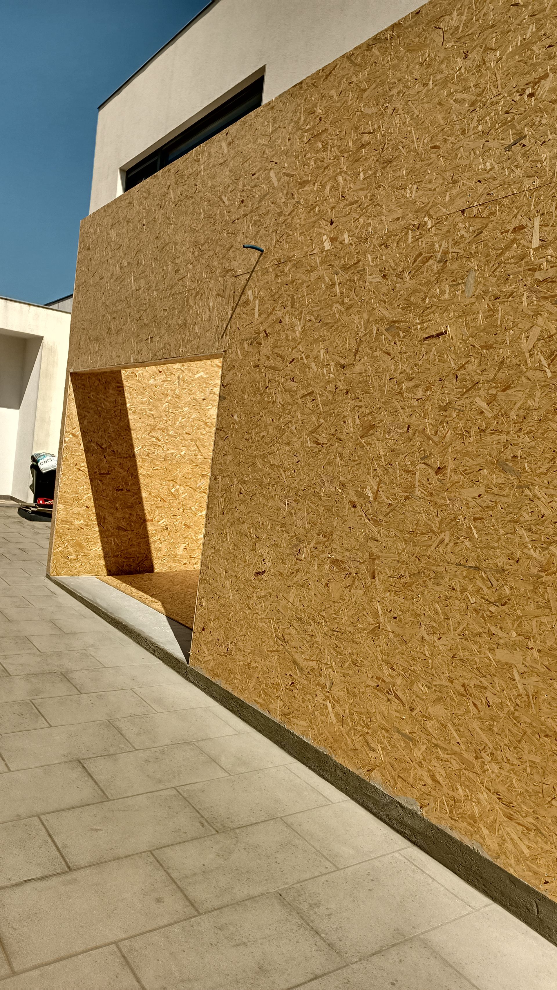 Vista exterior de um edifício com uma grande parede de aglomerado; uma abertura retangular projeta uma sombra na parede.