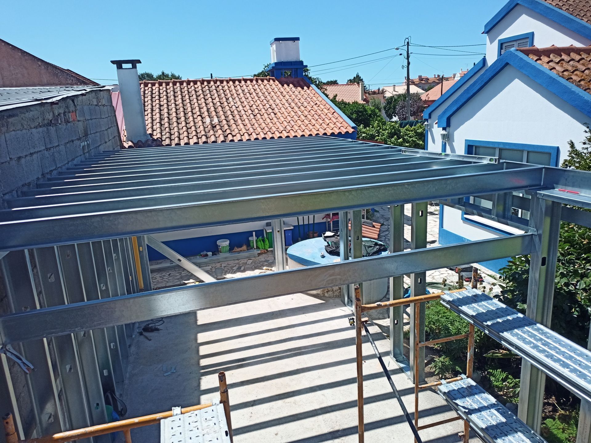 Estrutura metálica de um terraço em construção, com uma casa azul ao fundo.