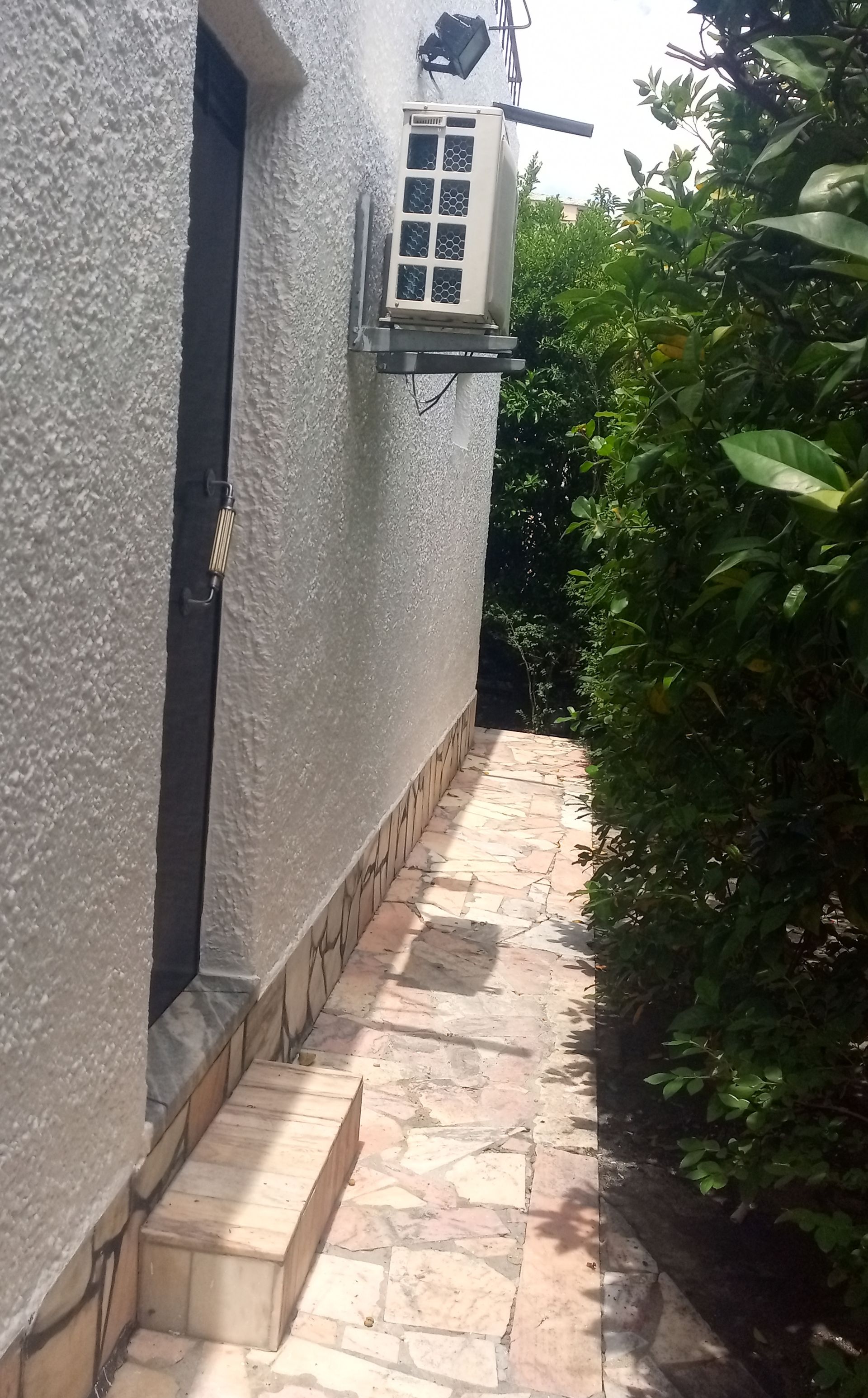 Caminho estreito de pedra ao lado de um prédio branco com ar condicionado. Arbustos verdes à direita.