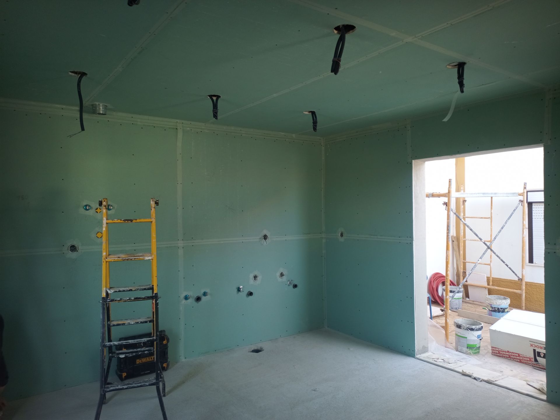 Sala interna com drywall verde em construção; fiação elétrica visível; escada presente.