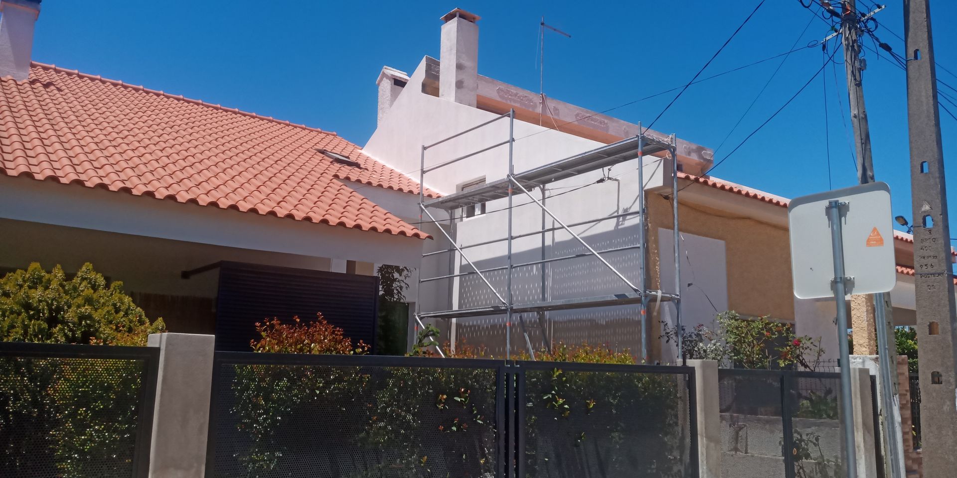 Casa com telhado de terracota, andaimes e portão de ferro forjado. Céu azul claro.