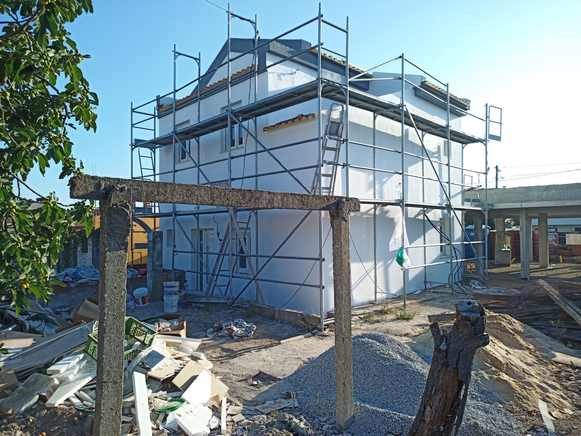 Prédio em construção, com andaimes, exterior branco, céu azul, materiais de construção em primeiro plano.