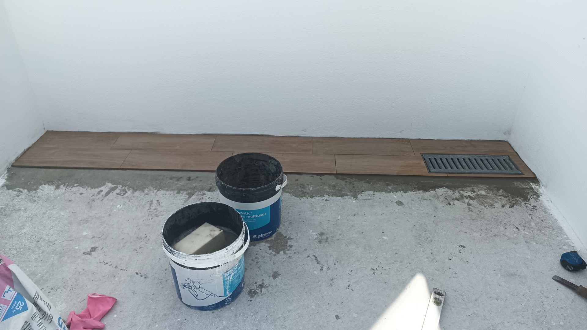 Dois baldes, tábuas de madeira e um ralo sobre um piso de concreto ao lado de paredes brancas, possivelmente em construção.