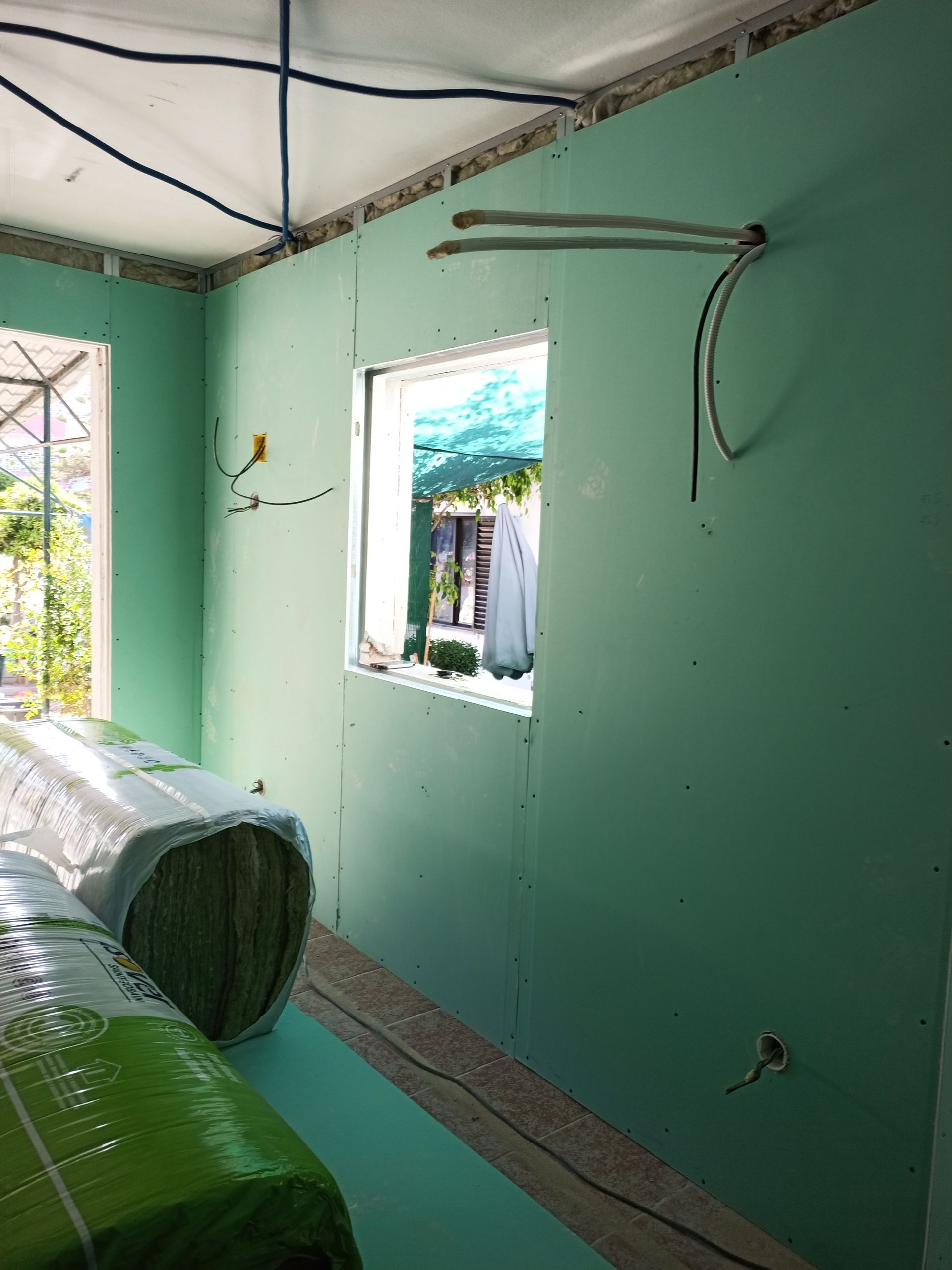 Interior de um cômodo em construção com drywall verde e fiação exposta. Rolos de isolamento estão no chão.