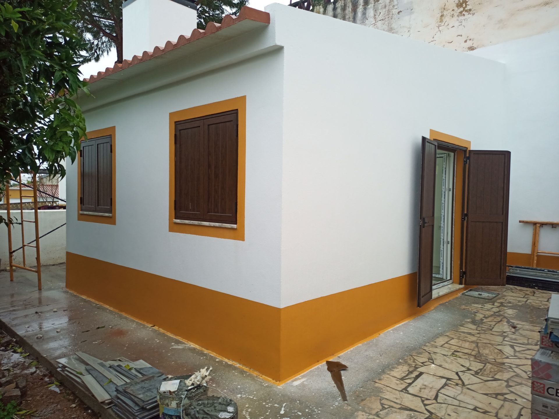 Prédio branco e amarelo com venezianas e detalhes em marrom. Pátio externo de concreto e tijolos encharcado pela chuva.