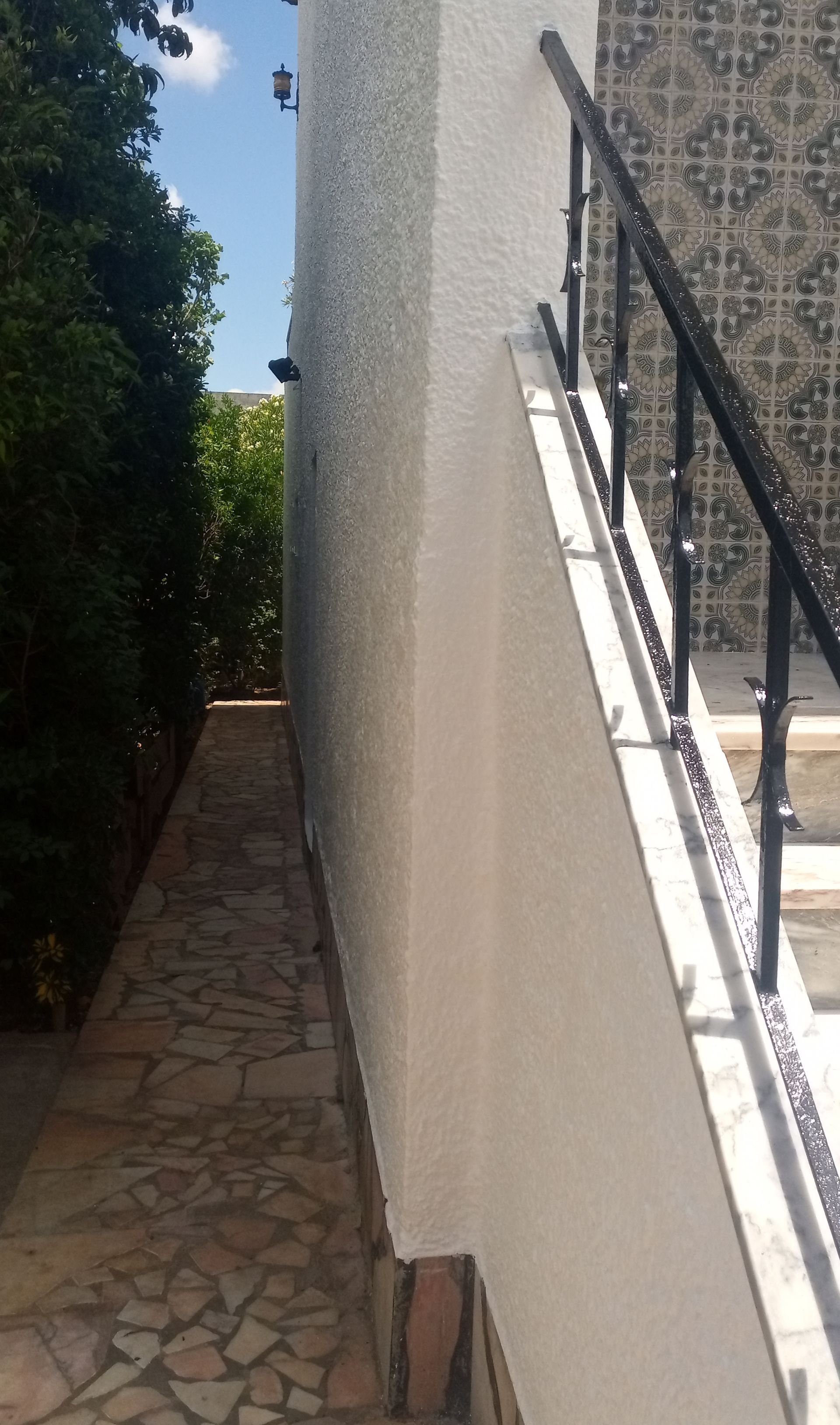 Caminho de pedra ao lado de uma parede branca texturizada e escada com corrimão preto. Árvores verdejantes à esquerda.