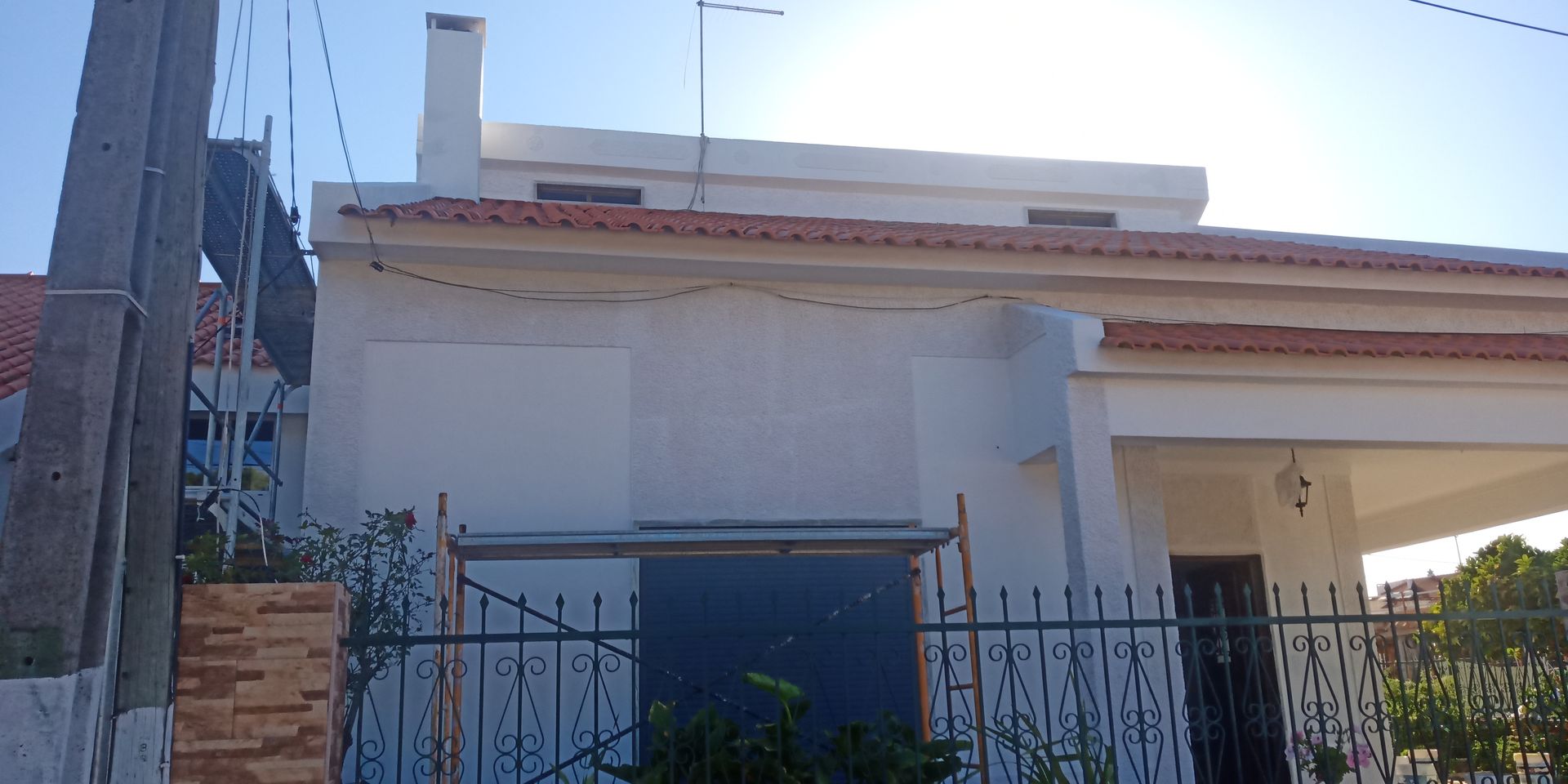 Casa branca com telhado de telha vermelha, chaminé, cerca e fios de energia contra um céu azul.