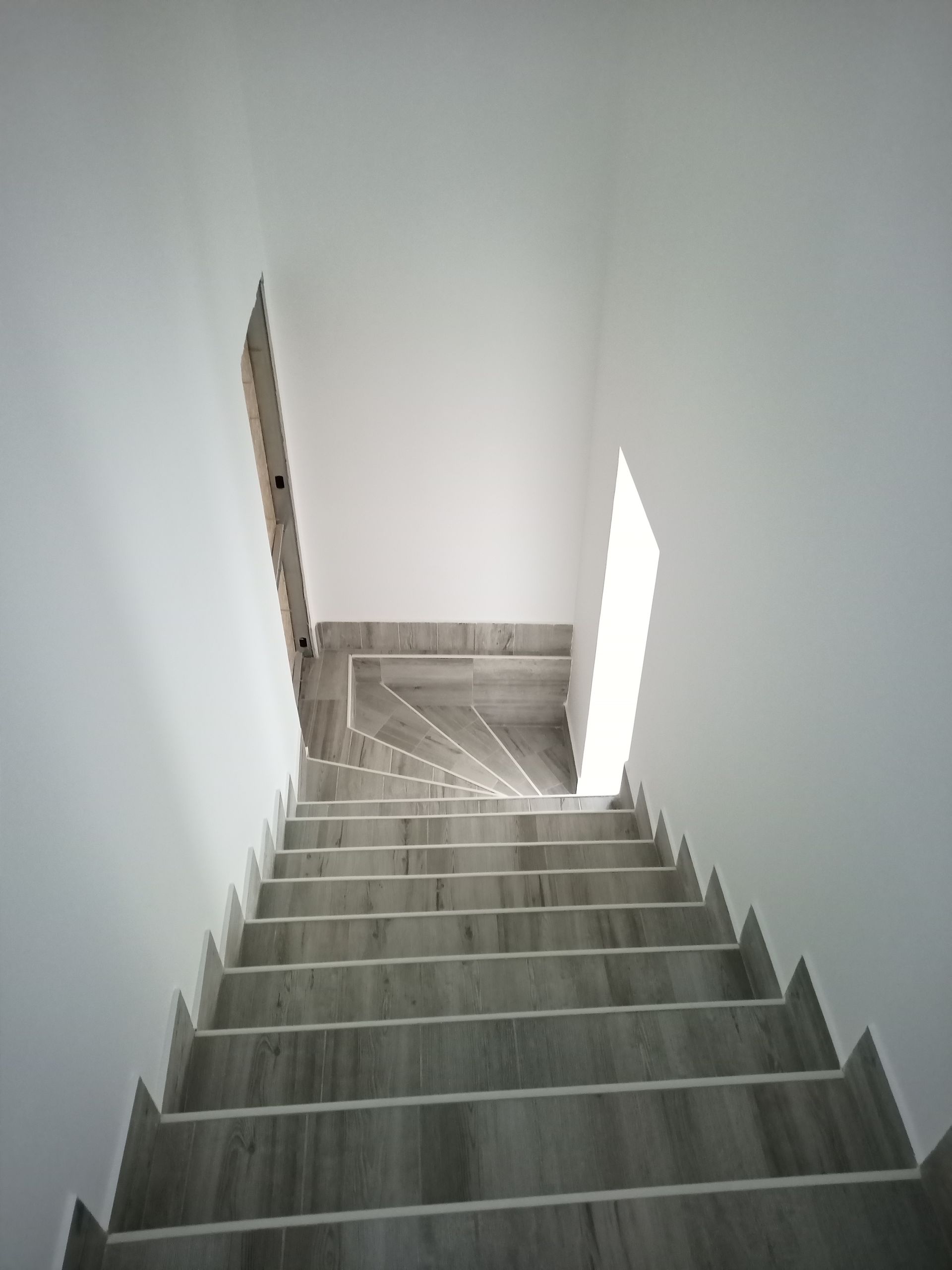 Escadaria que sobe com degraus cinzentos e paredes brancas; a luz natural brilha por cima.