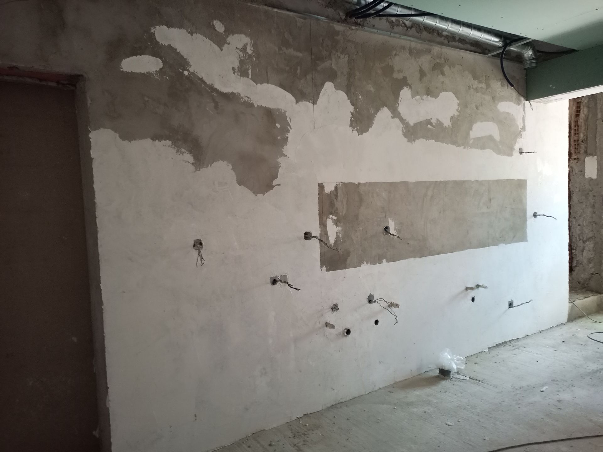 Parede de obra com tomadas elétricas, reboco inacabado e fios expostos. Uma seção retangular está sem reboco.