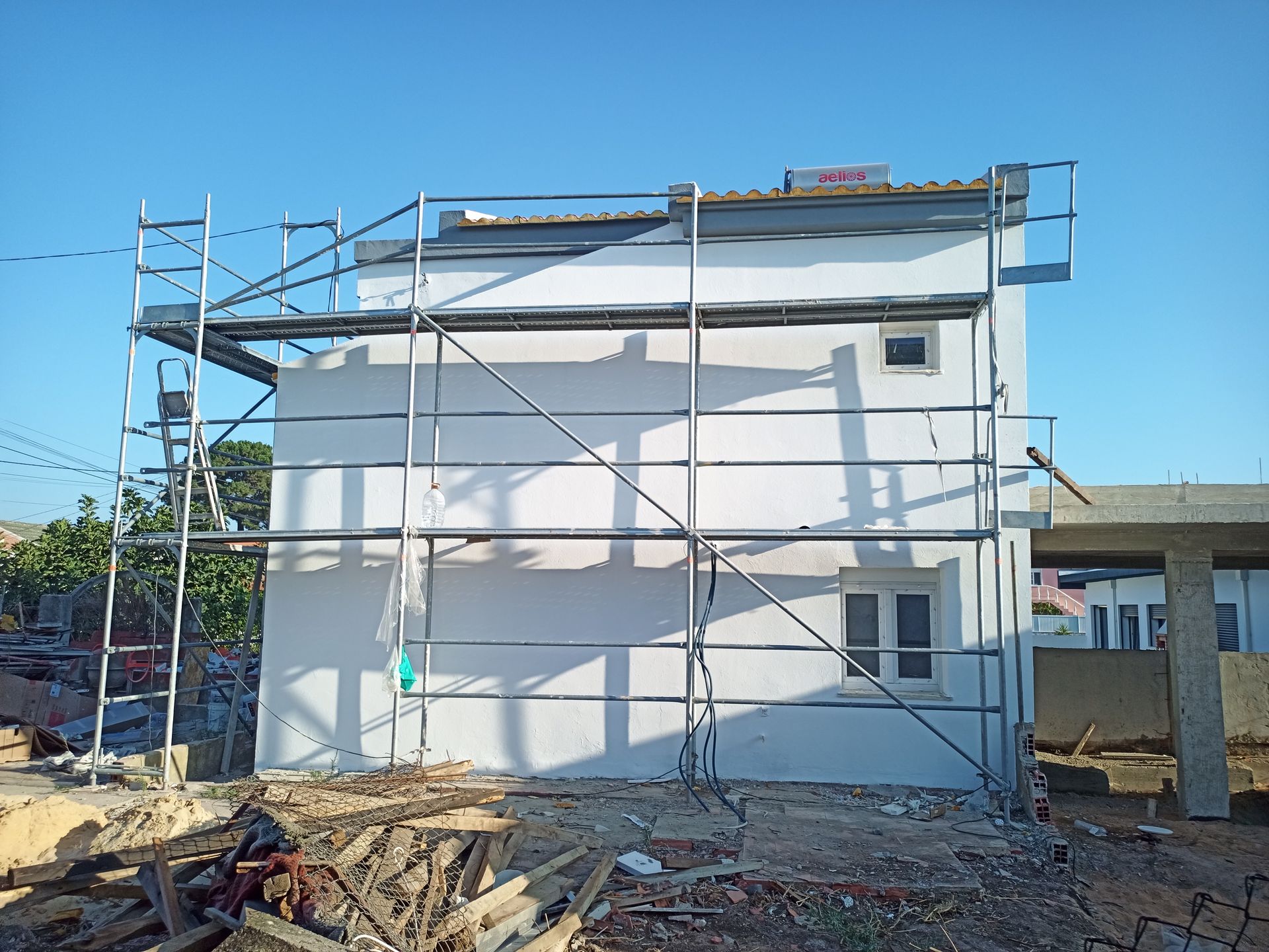 Em um dia ensolarado, andaimes cercam um prédio de dois andares com paredes brancas, em construção.